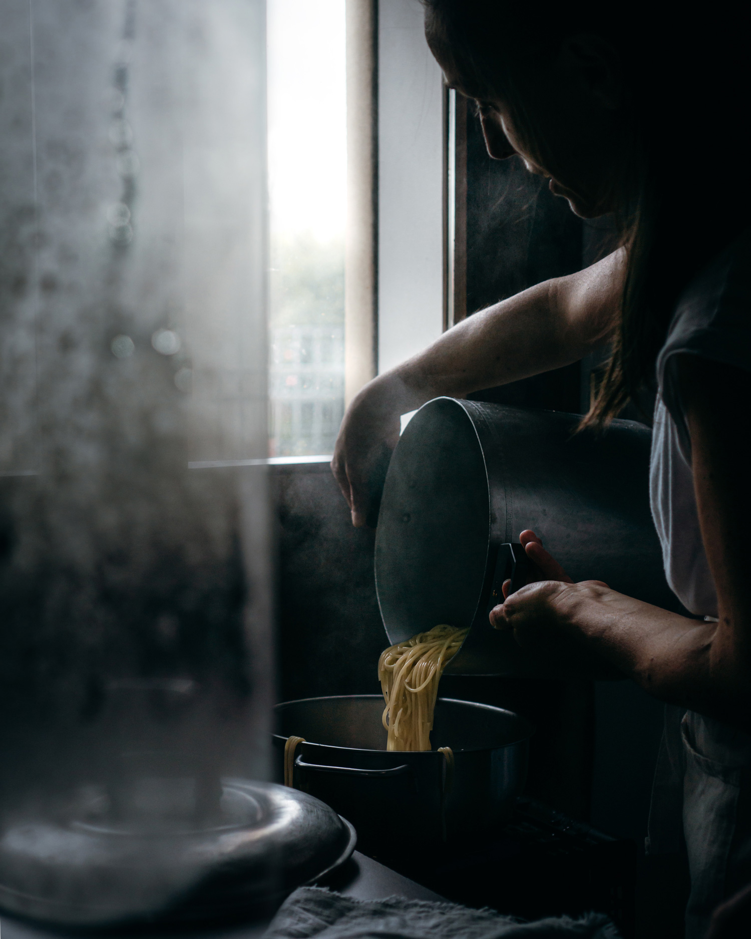 Elisa De Cecchi stands near a window, draining spaghetti from a saucepan into a strainer.
