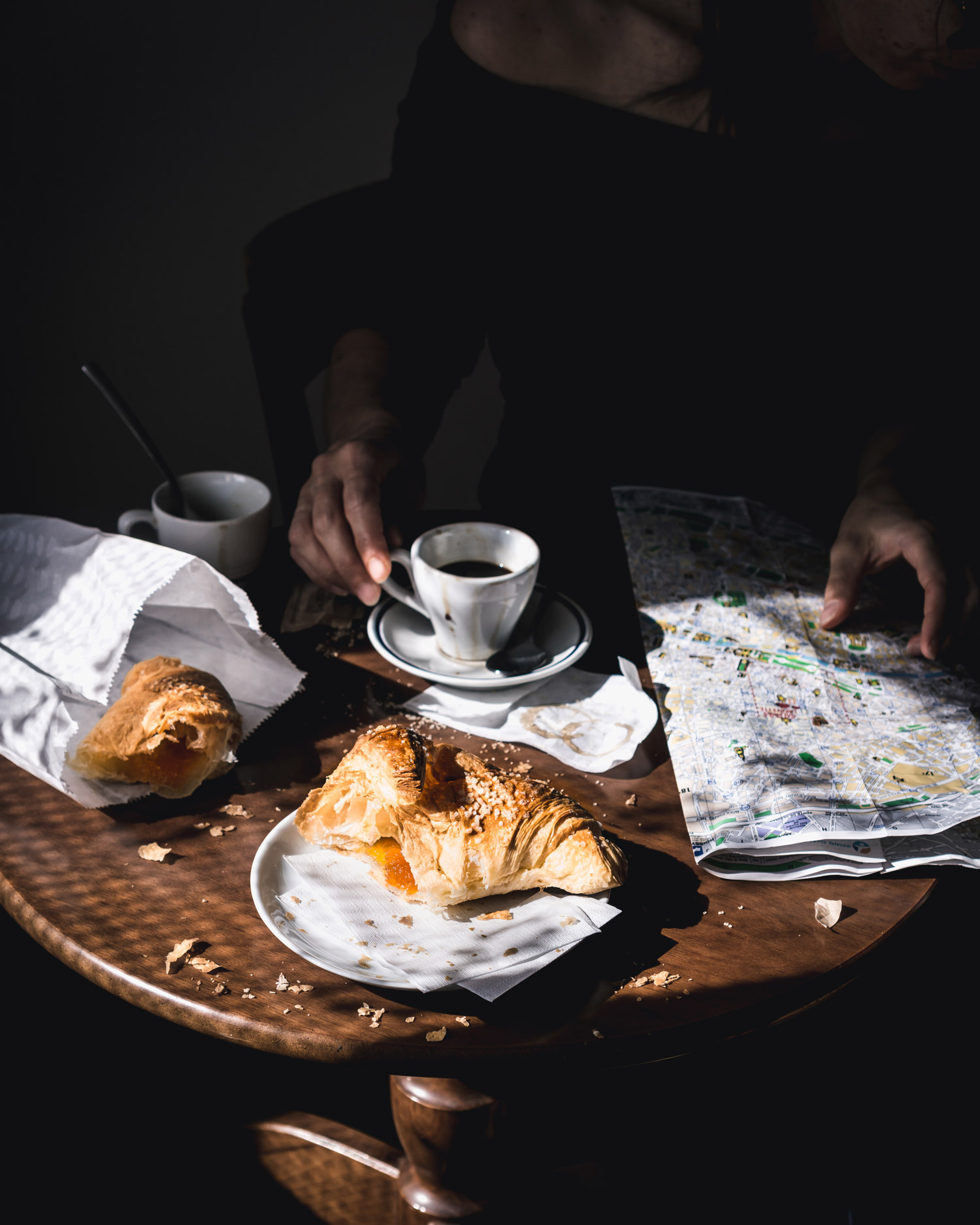 Two croissants, two cups of coffee and a map on a table, with hands on a cup and the map.