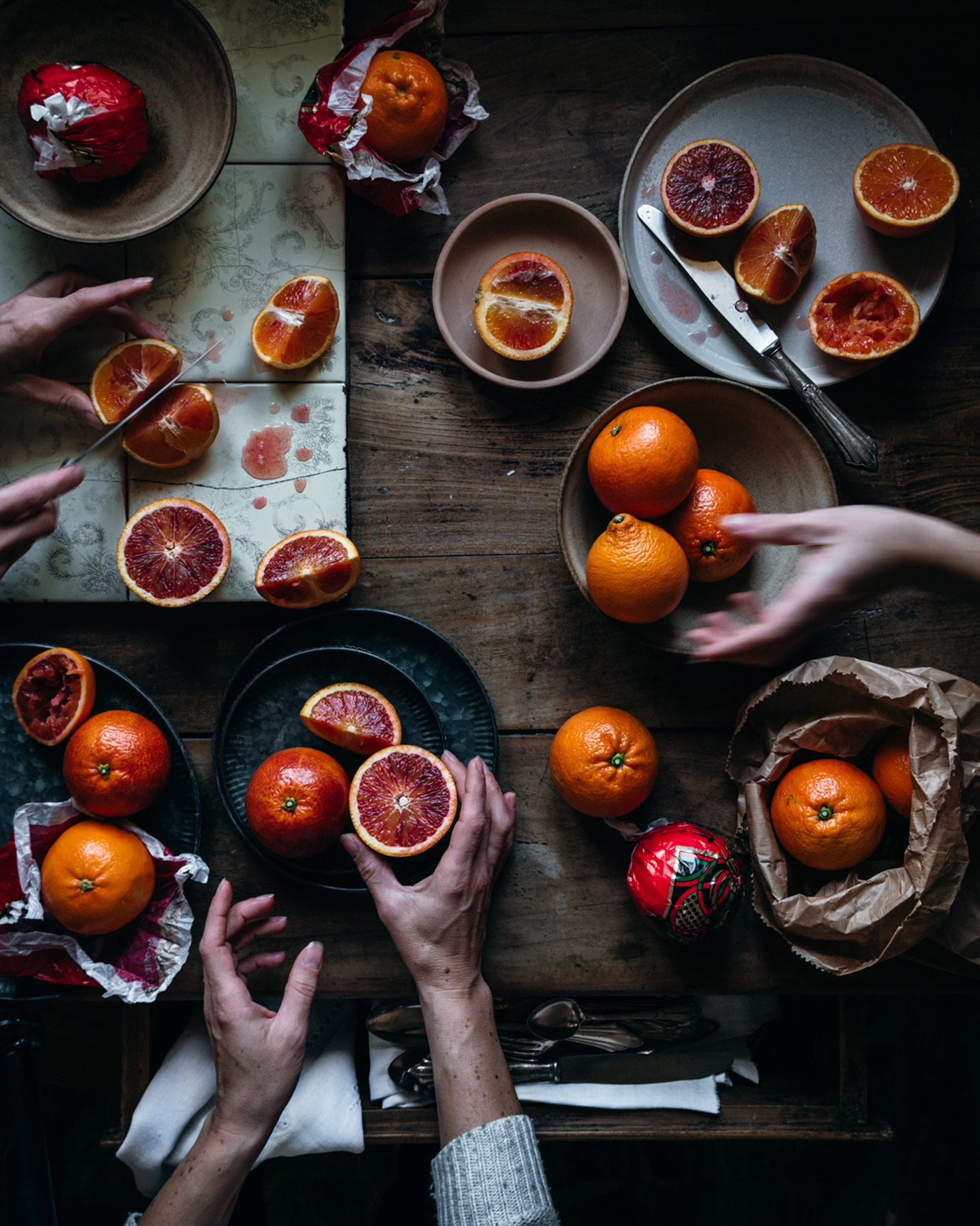 A table full of blood oranges, some cut in half or segments, with five hands chopping and holding them.
