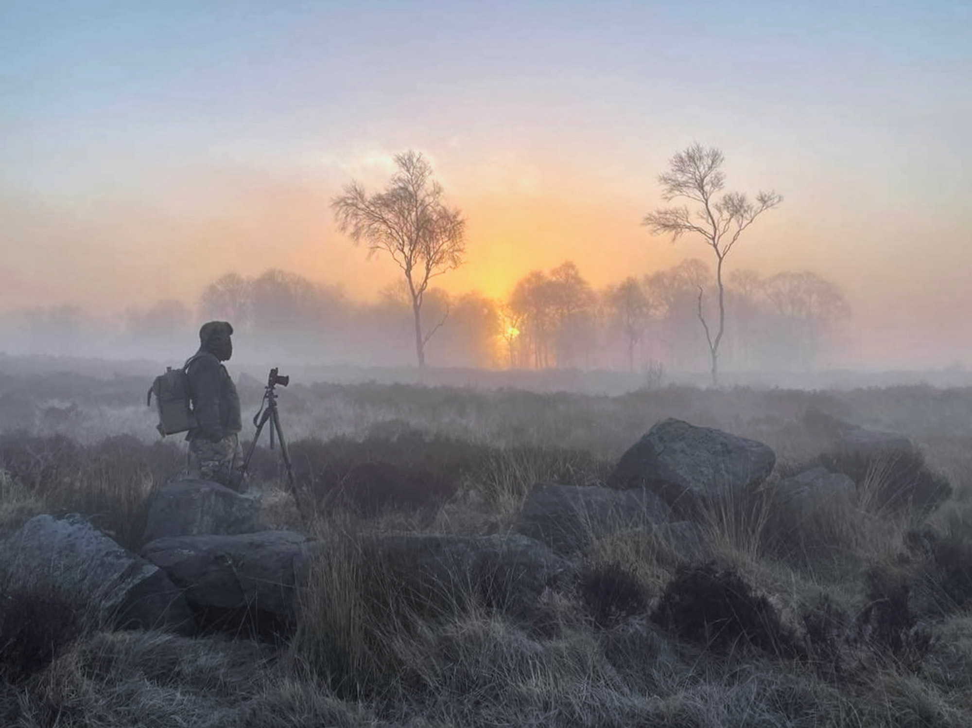 Chris Nowell stands in a field with a camera, with trees and the sun in the background.