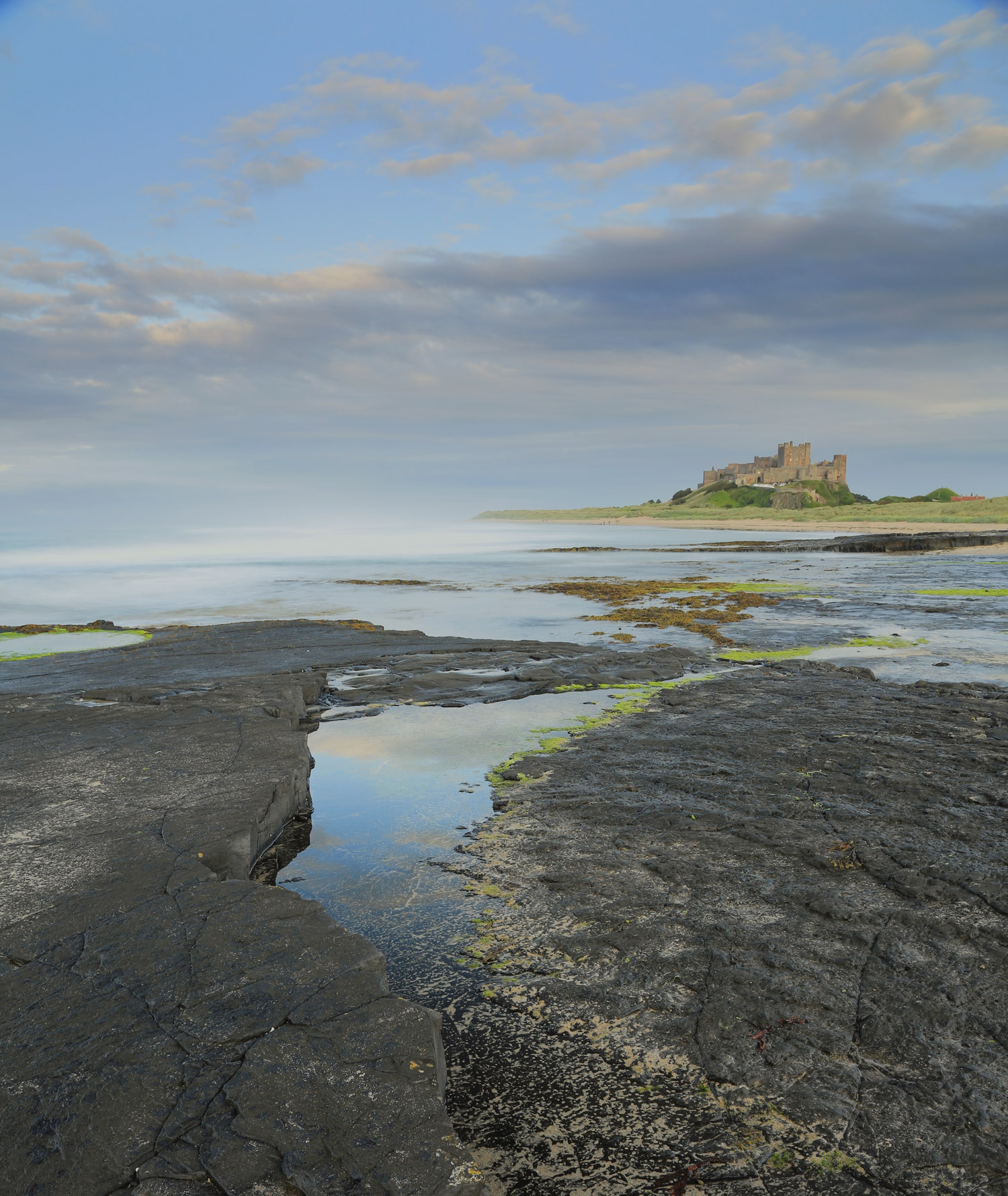 Rocks with water on in front of Bamburgh Castle on a hill in the background.