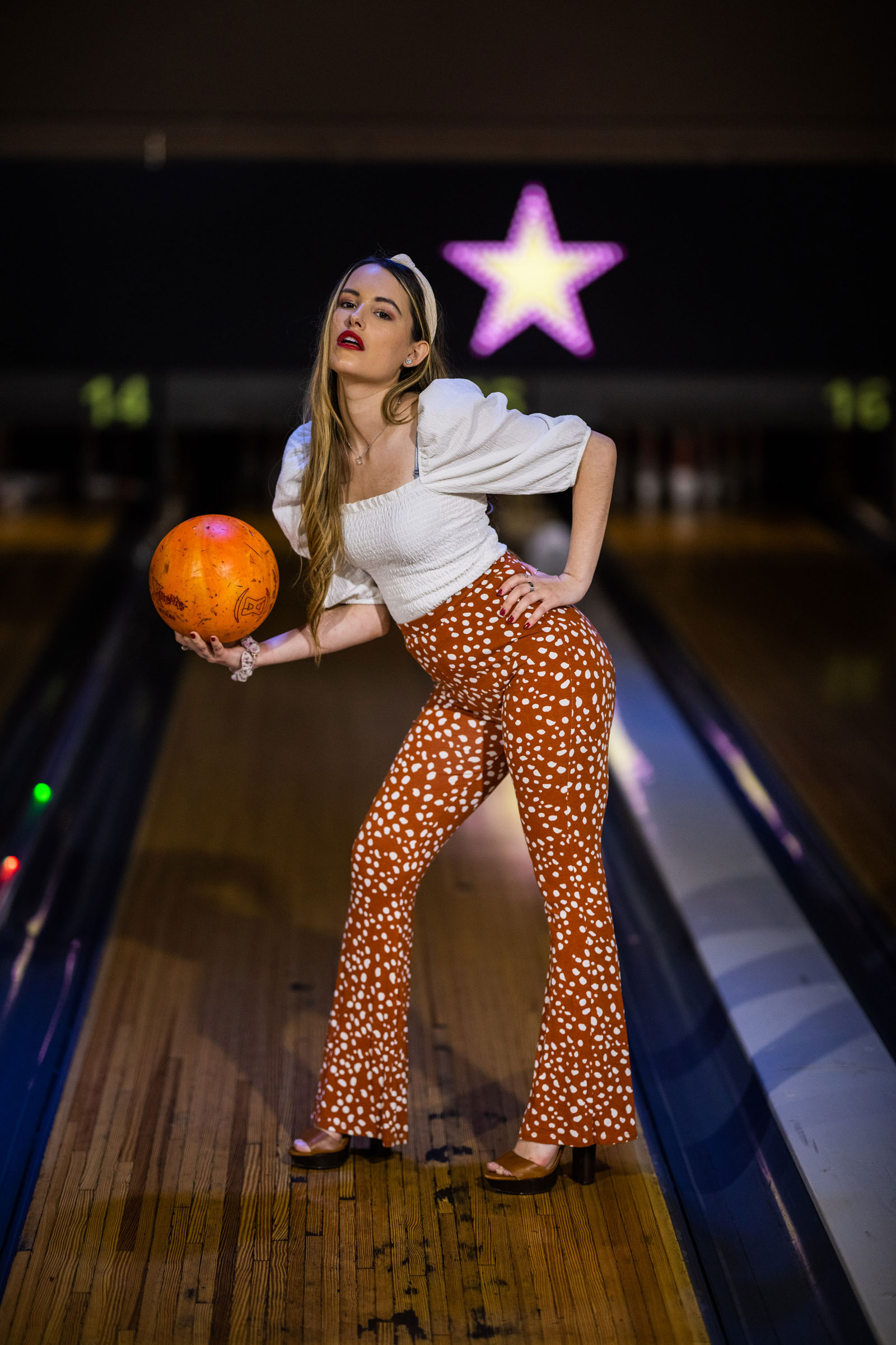 A model wearing orange trousers and a white top stands holding a bowling ball in a bowling alley. Shot with Canon EOS R5 + Canon RF 85mm f/1.2 L