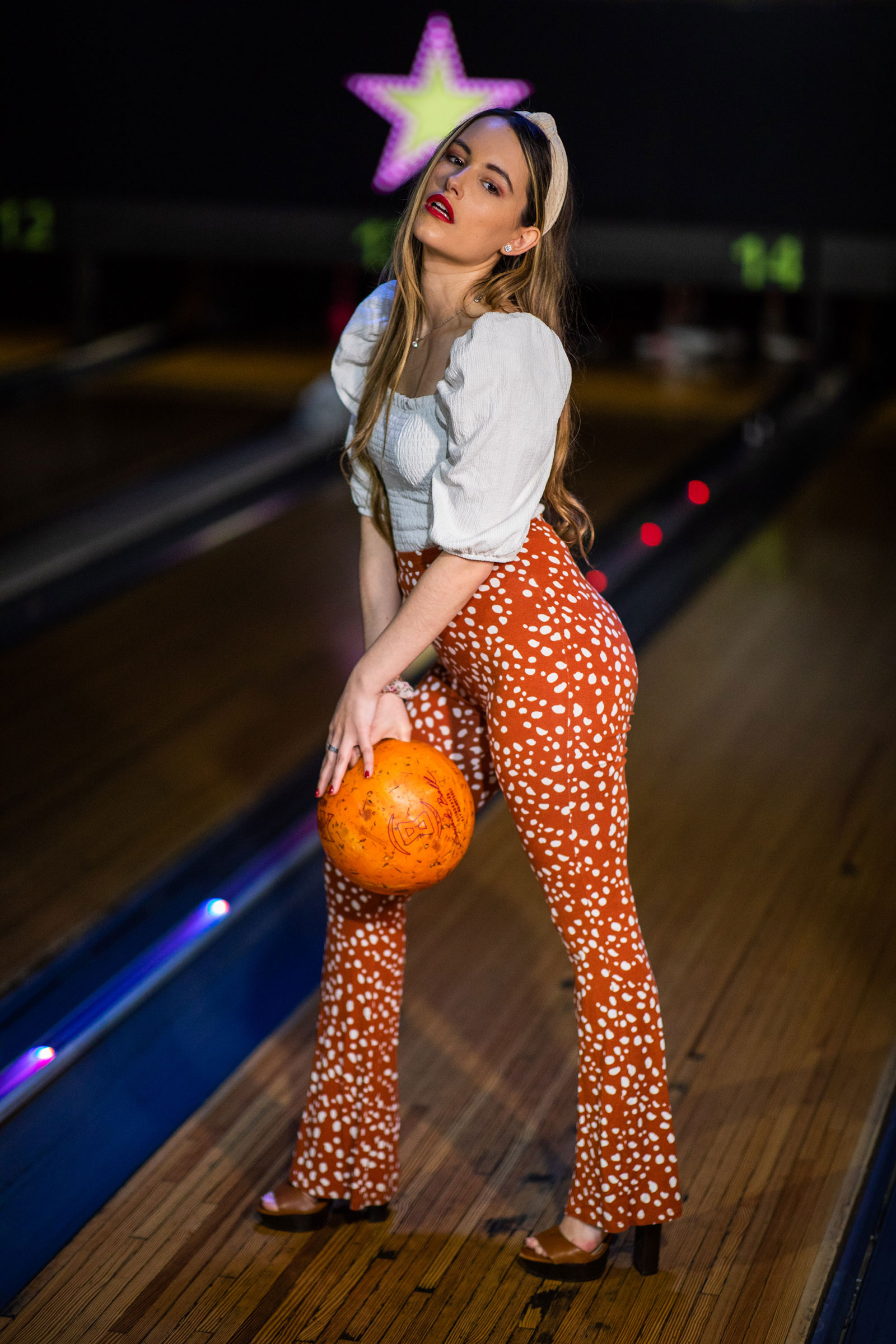 A model wearing orange trousers and a white top stands holding a bowling ball in a bowling alley. Shot with Canon EOS 5D Mark IV + Canon EF 85mm f/1.4 L IS