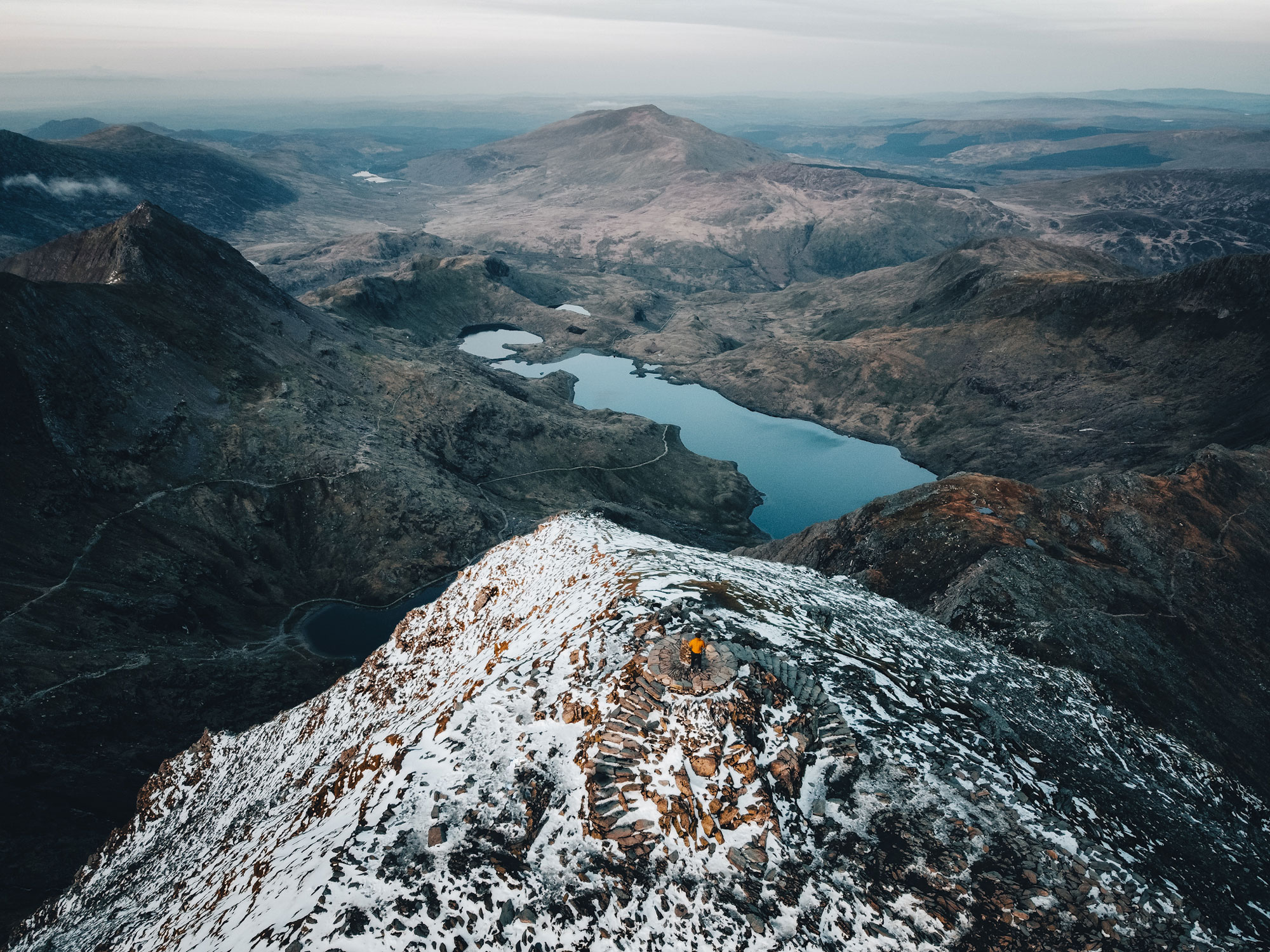 Une vue aérienne d'une chaîne de montagnes avec un lac au fond de la vallée. Un personnage portant une veste orange se tient au sommet d'un pic.