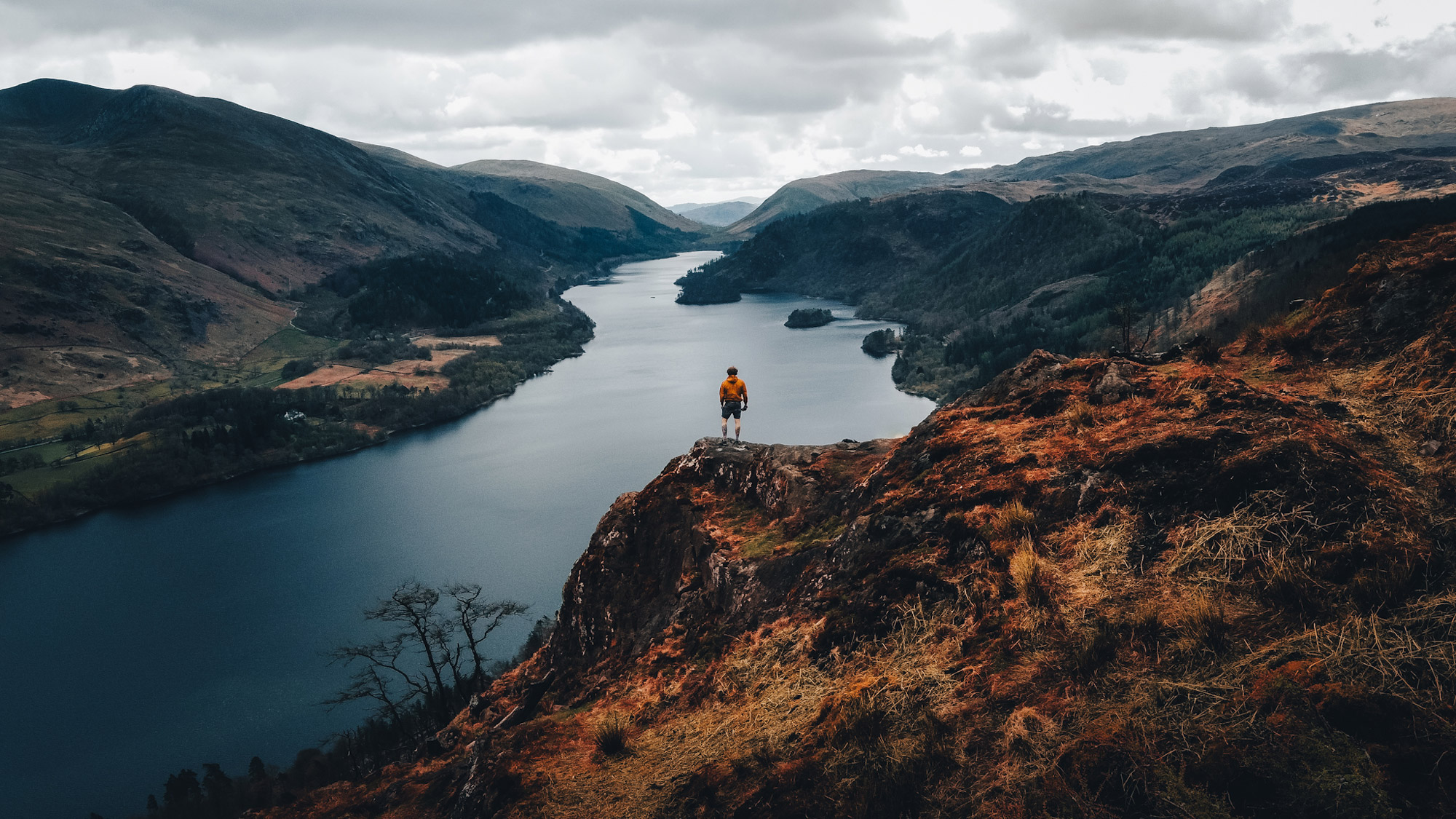 Une chaîne de montagnes traversée par une rivière. Un personnage portant une veste orange se tient au sommet d'un pic.