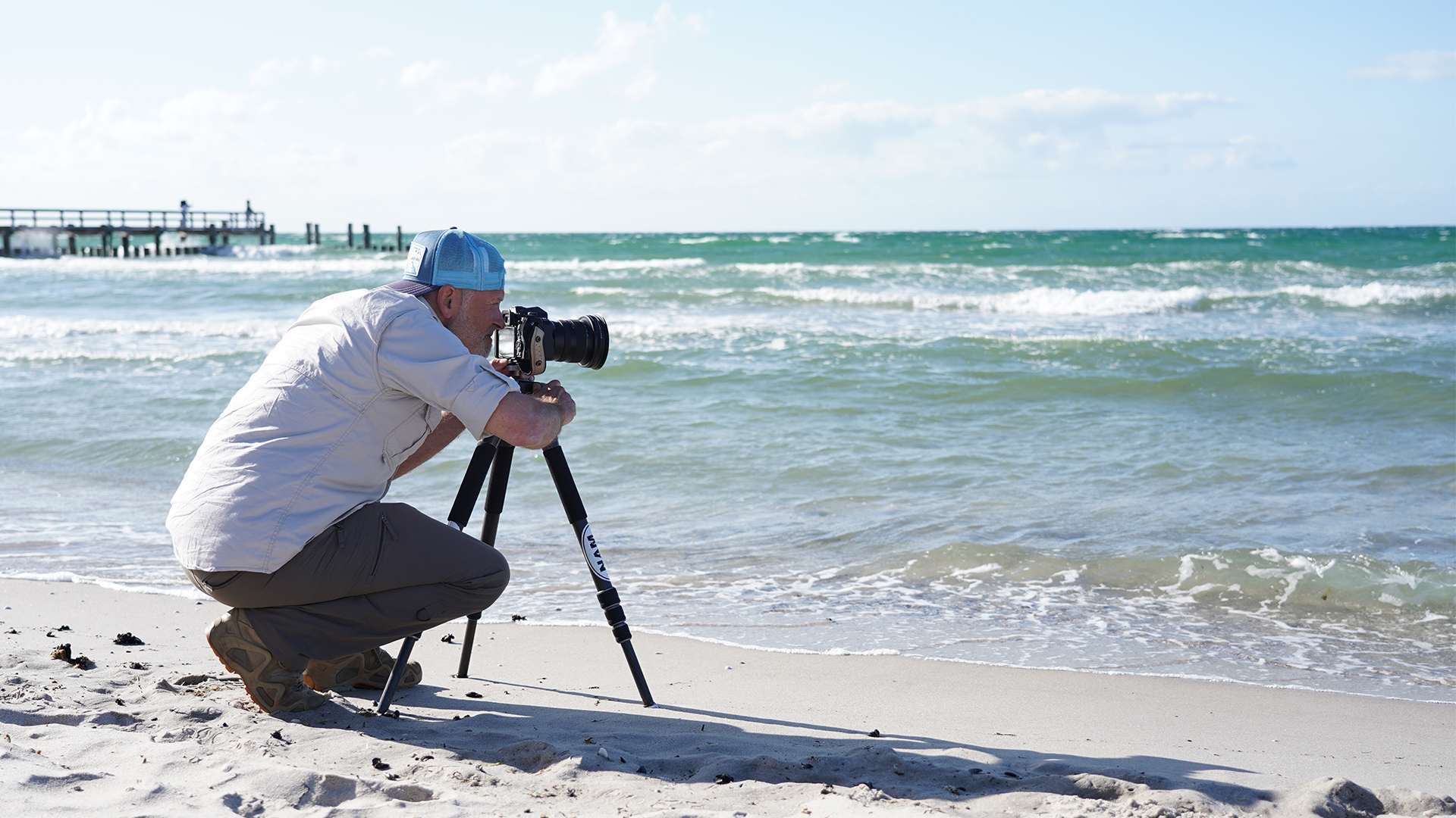 Ein Mann kniet vor dem Meer, während er durch eine Kamera schaut 
