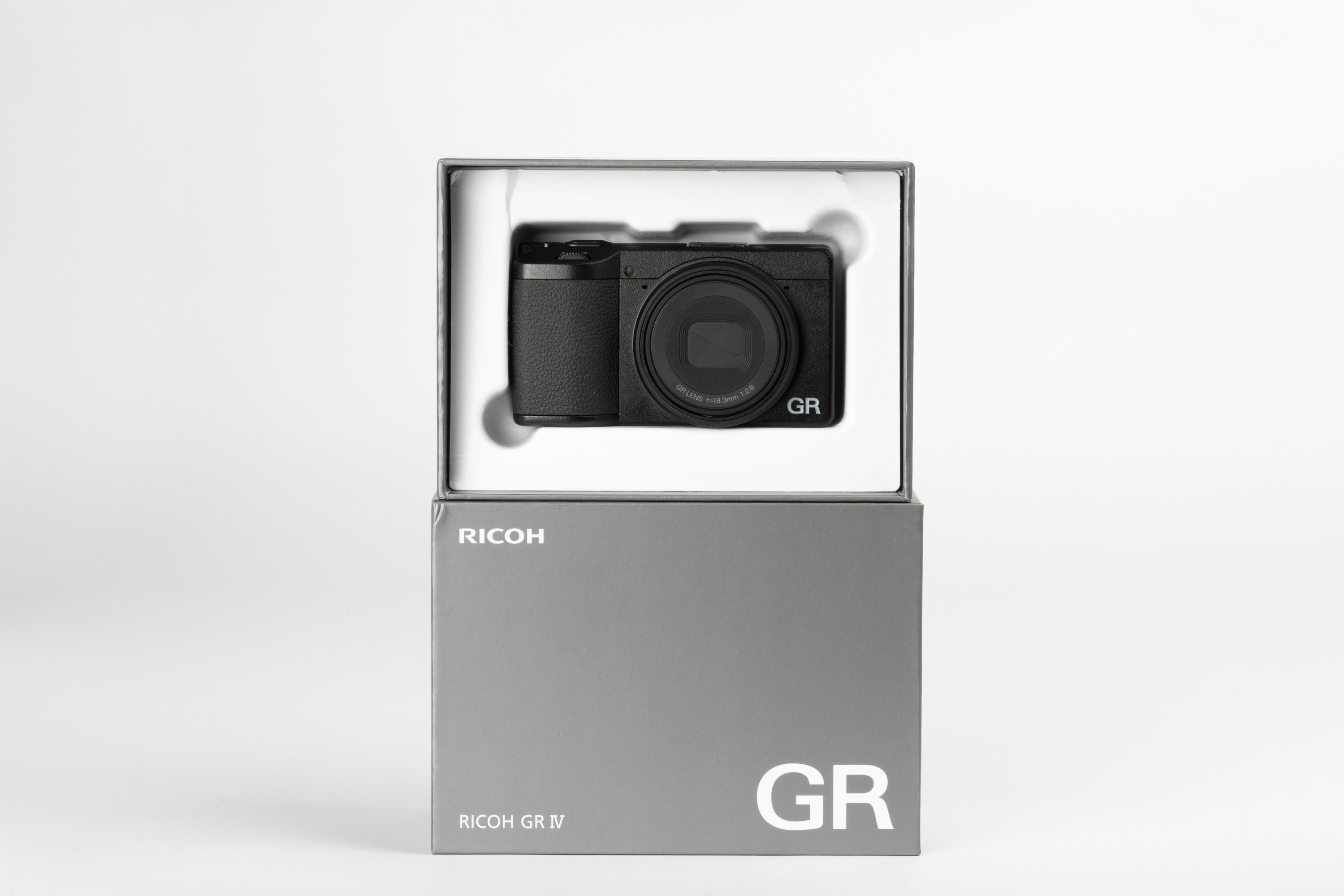 Eine Ricoh GR IV in ihrer Originalverpackung auf cremefarbenem Hintergrund. Der Umkarton ist grau, die Innenverpackung weiß.