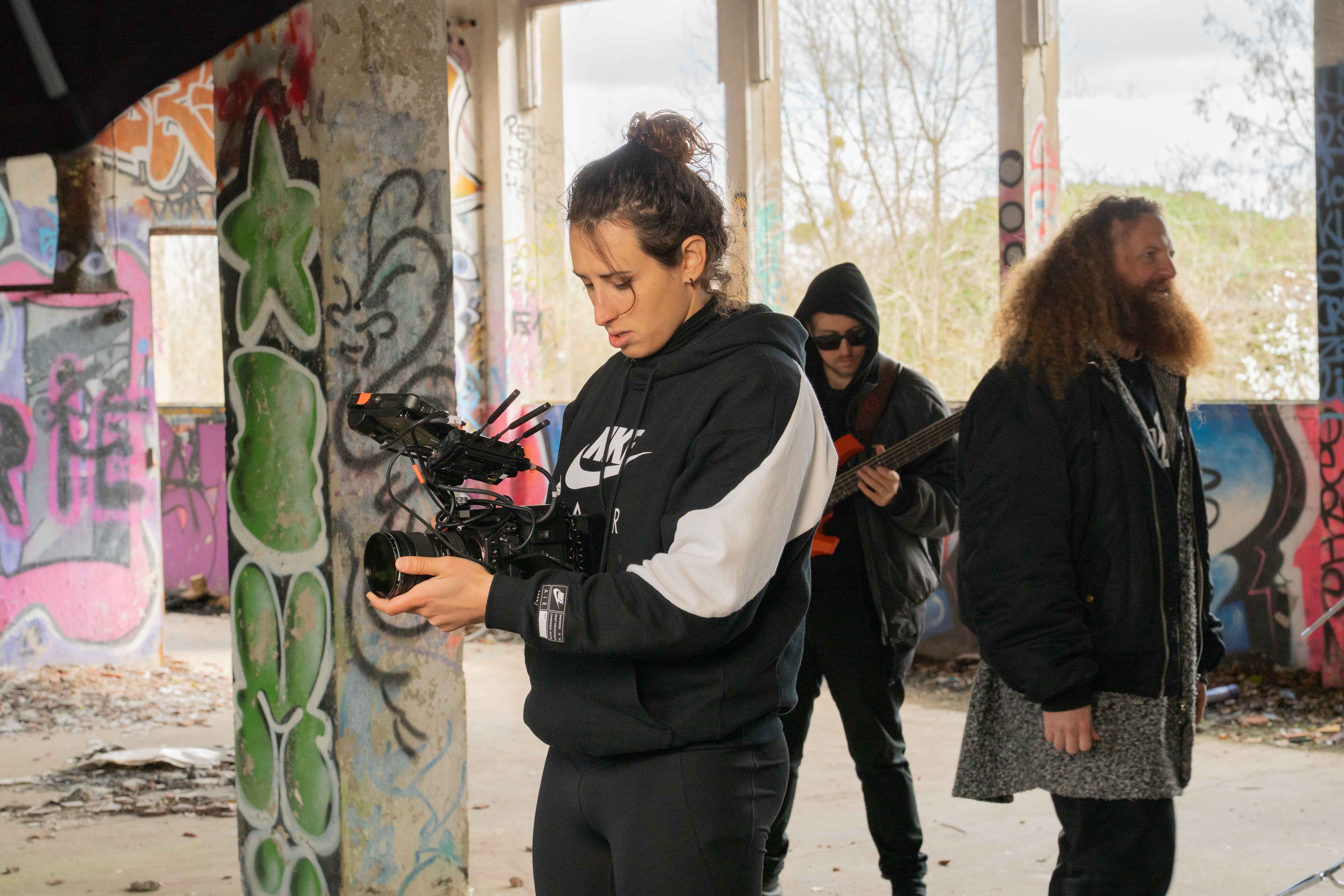 Lena Hempel adjusts a professional camera rig inside a graffiti-covered building while two band members stand in the background, one holding a bass guitar.