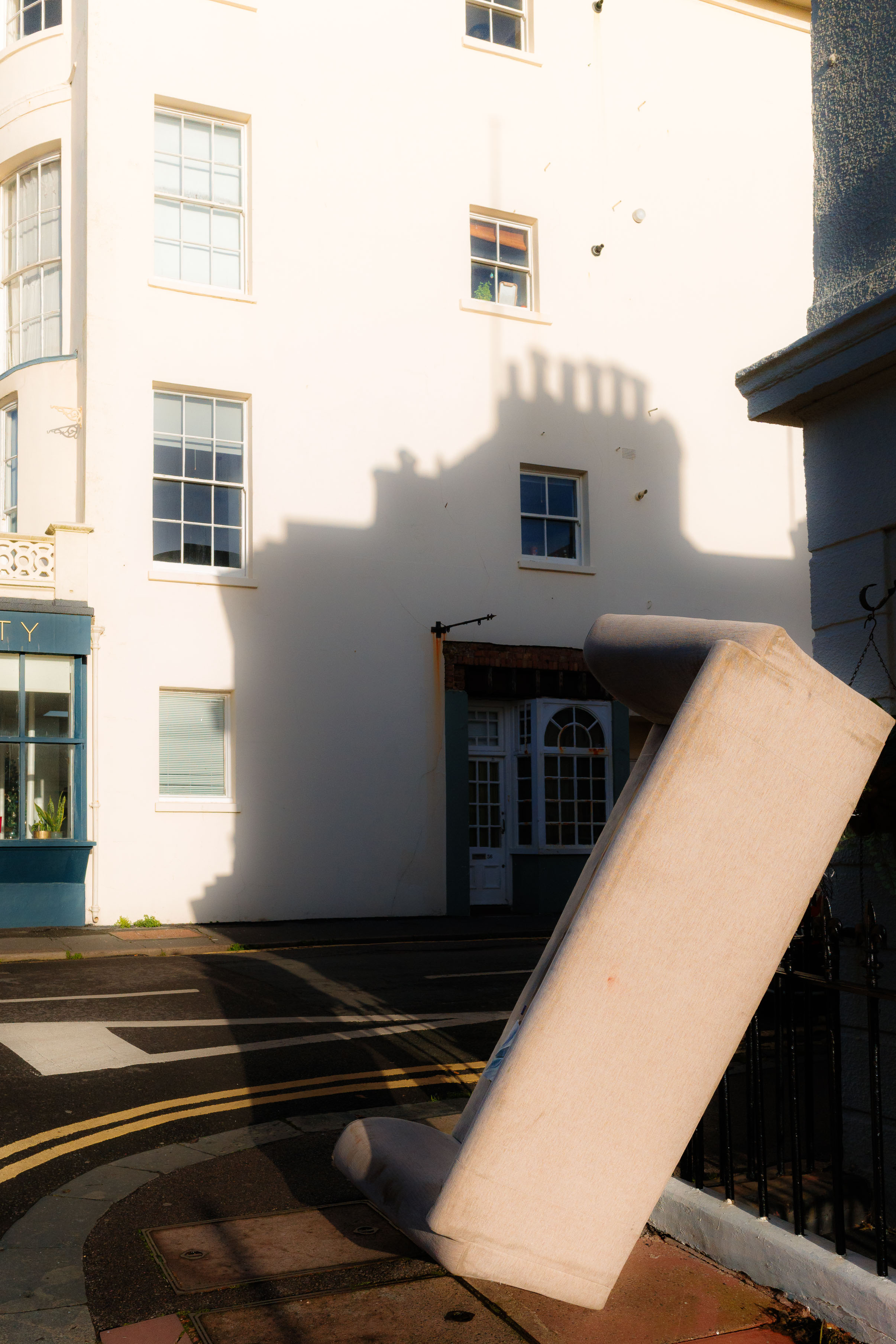 Streetaufnahme, die ein weggeworfenes Sofa zeigt, das an einem Geländer lehnt. Im Hintergrund, starkes Sonnenlicht und architektonische Schatten an einem weißen Gebäude. Aufgenommen mit einer Ricoh GR IV.