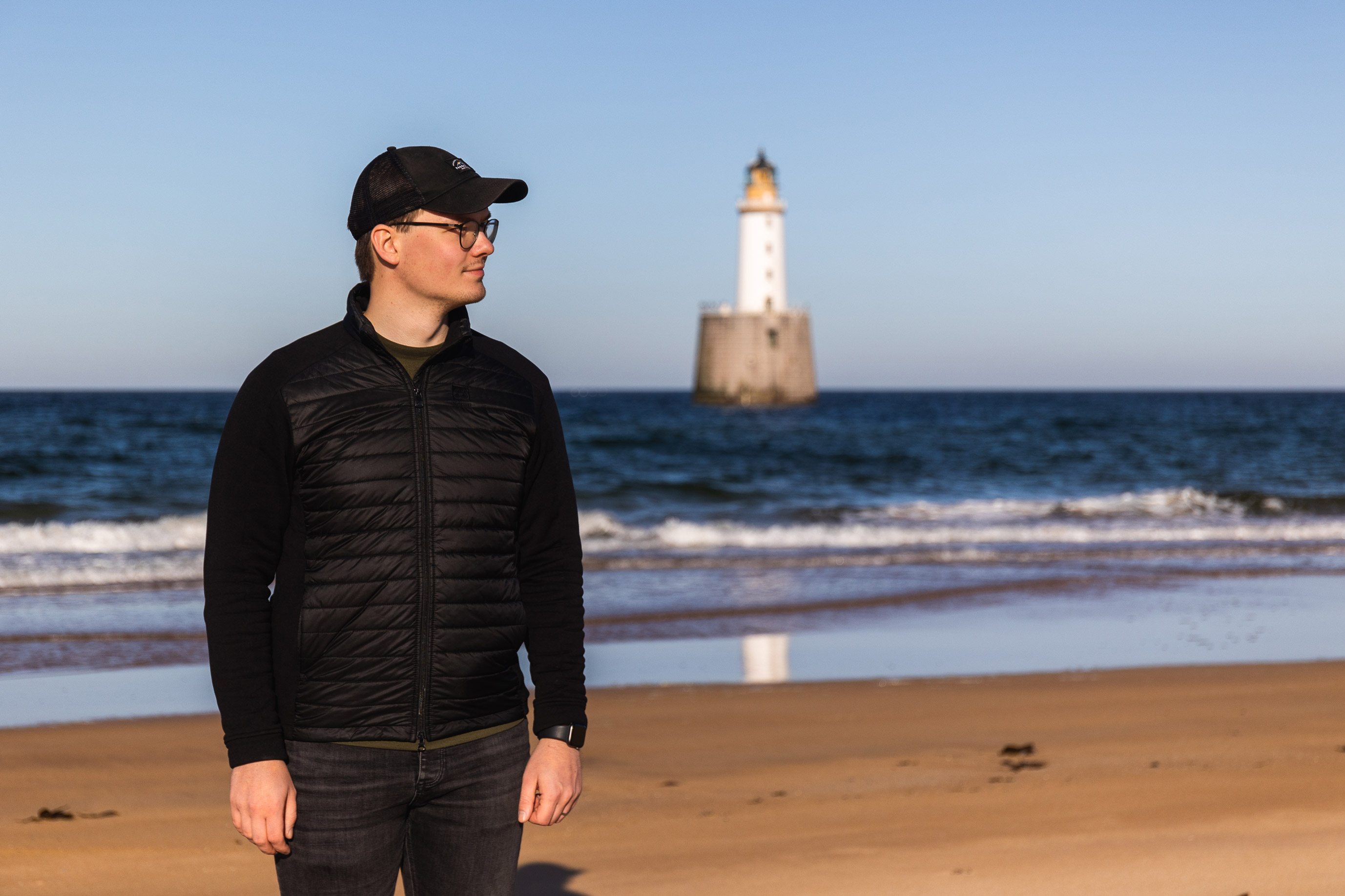 a man wearing a hat standing on a beach with the sea and a lighthouse in the background