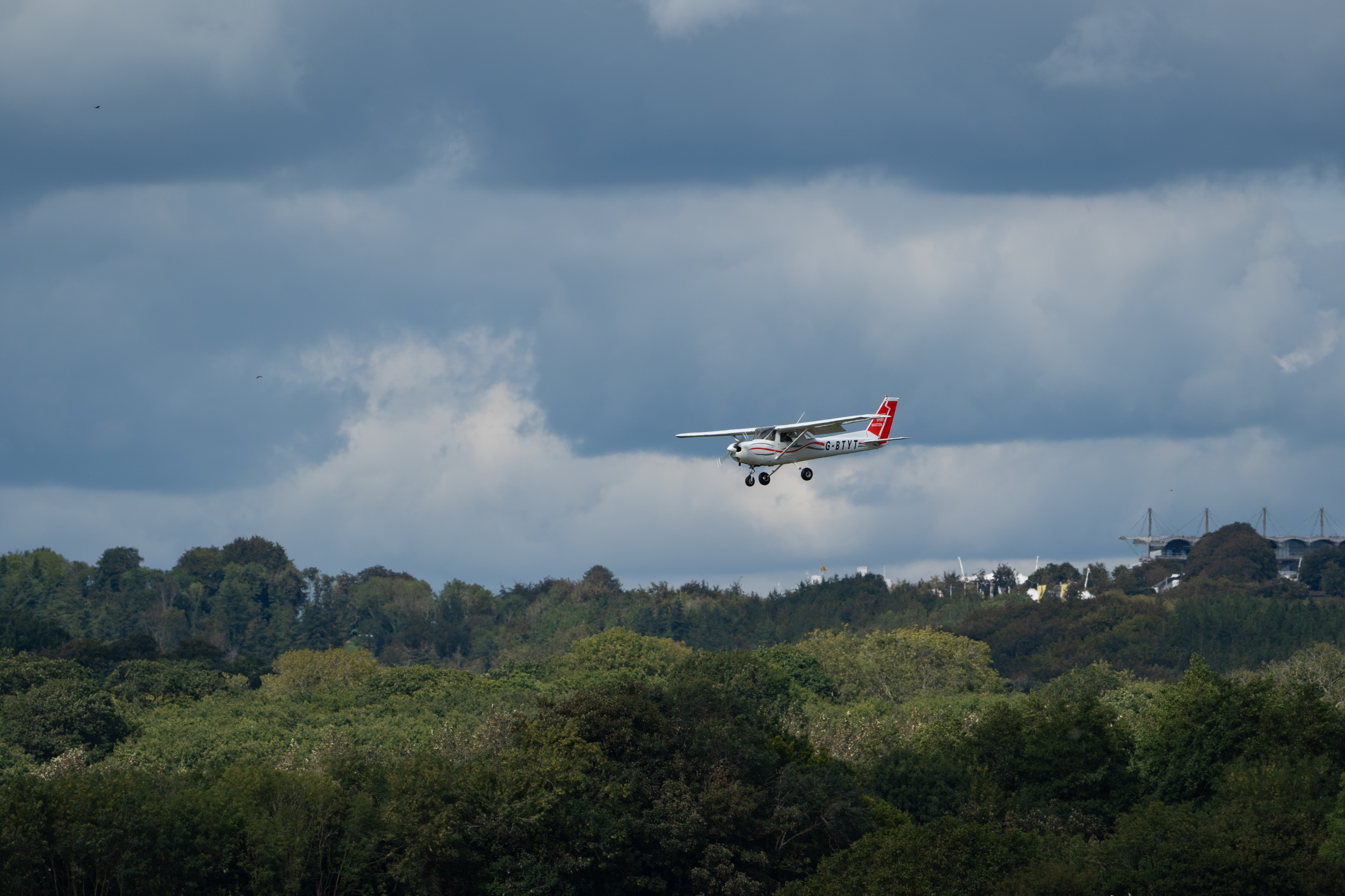 A small glider plane flies through the sky, with thick grey clouds in the background. There is a forest beneath it, with the different shades of green leaves and clouds creating a layered effect.