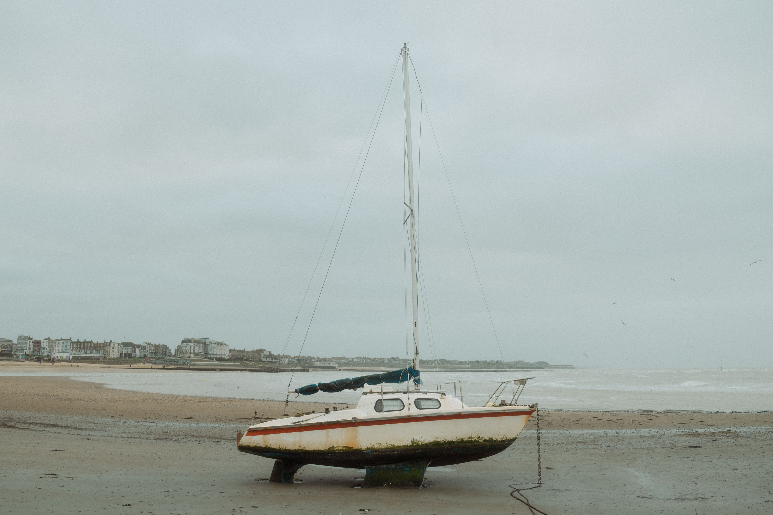 Ein kleines Segelboot, das bei Ebbe auf dem nassen Sand am Strand von Margate ruht, fotografiert an einem bewölkten Tag mit ruhiger See und entfernten Küstengebäuden im Hintergrund. Aufgenommen von Connor Redmond mit einer Sony RX1R III.