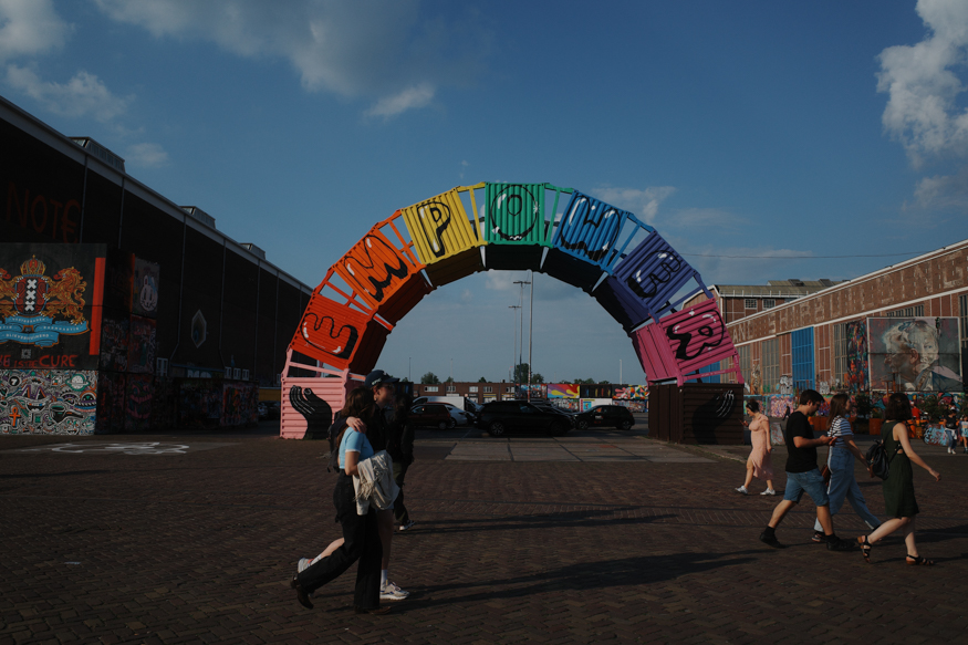 Een kleurrijk straatbeeld op het NDSM-terrein, waar voorbijgangers onder de iconische Empower-boog lopen in helder middaglicht.