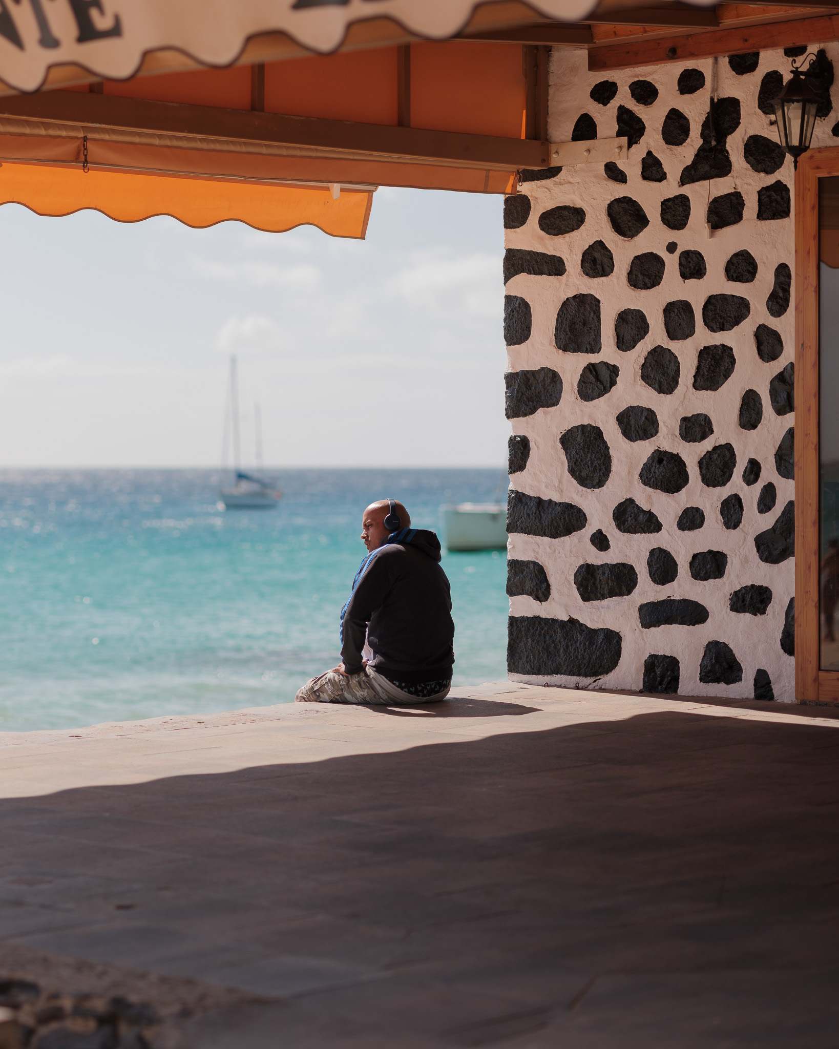 Eine Person mit Kopfhörern blickt nachdenklich auf das Meer, während sie am Rande einer Terrasse sitzt. Boote im Hintergrund. Von Connor Redmond.