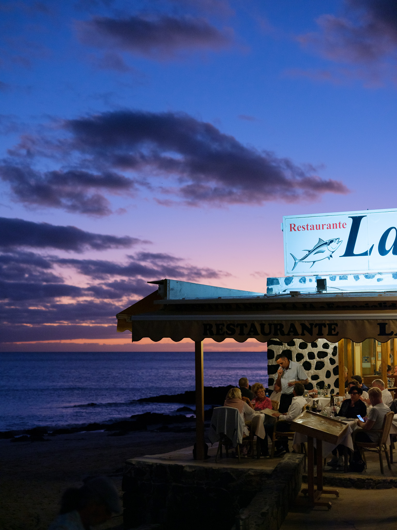 Restaurant next to the sea at sunset with people dining outside. By Connor Redmond.