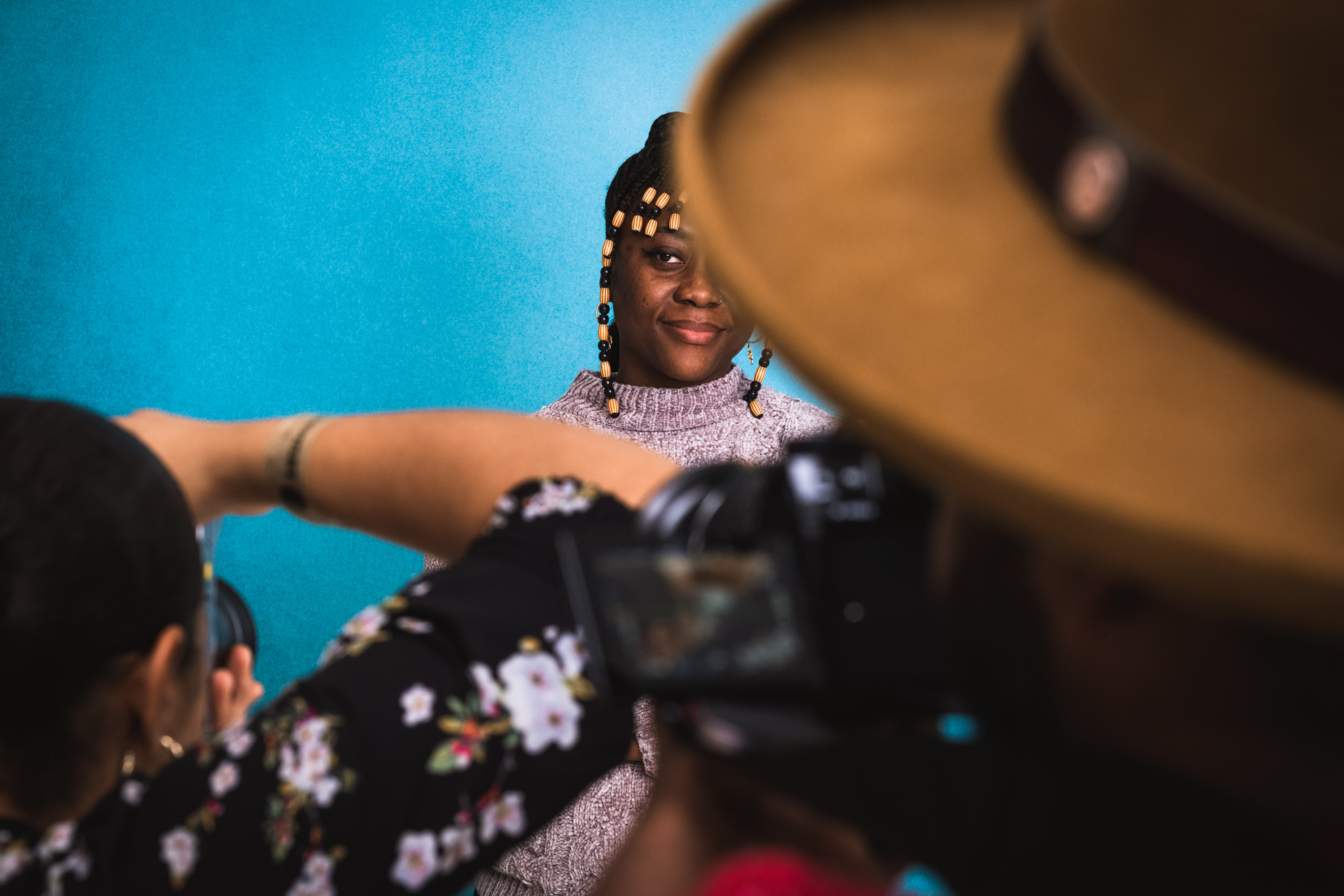 Two photographers capturing the portrait of a model, with braided hair wooden brown and black beads, wearing a purple sweater, against a light blue background, shot with a Fujifilm X-T5
