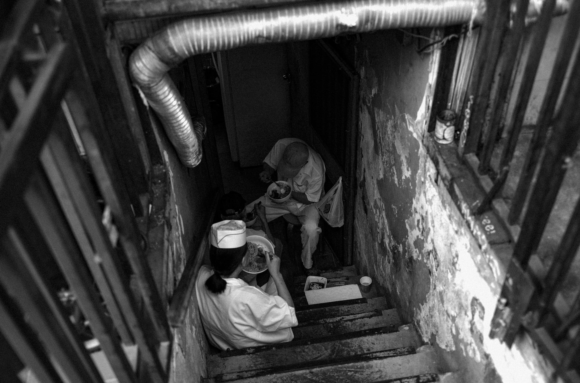 A black-and-white photo of two people sitting and eating at the bottom of a staircase