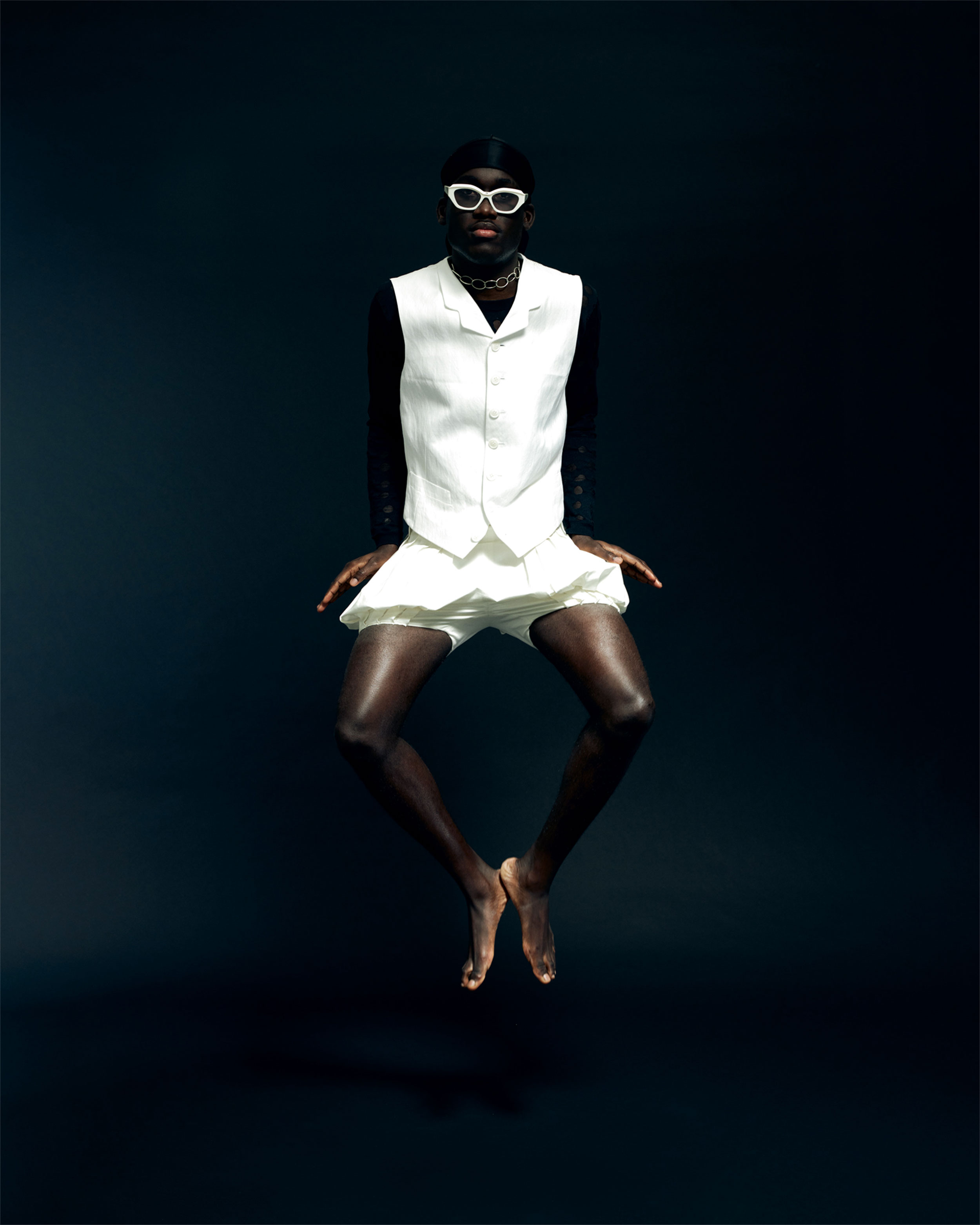 Homme noir portant des vêtements blancs et des lunettes de soleil blanches sur un fond sombre, semblant flotter avec ses jambes pliées. Photo par Lisa Marie Asubonteng