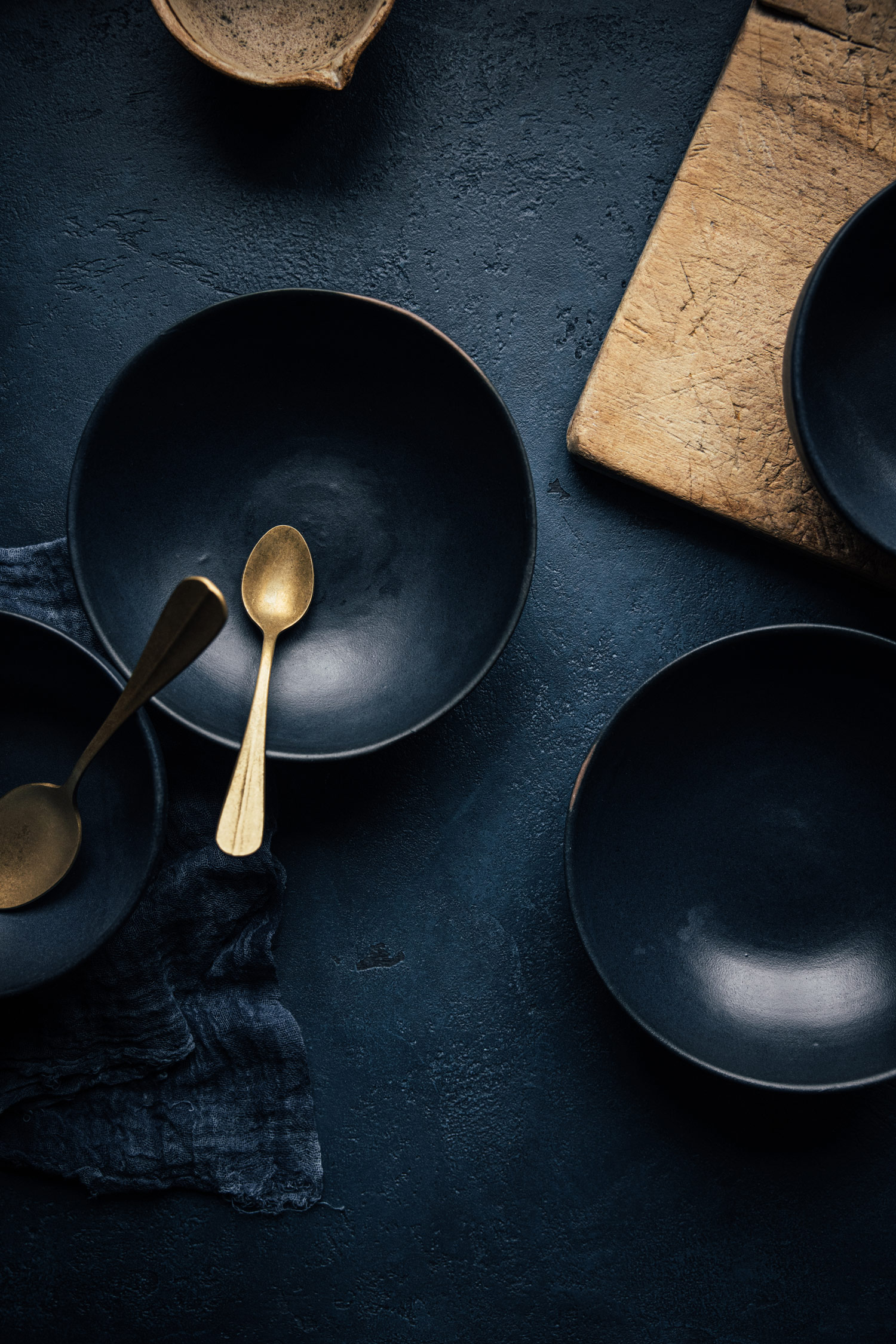 A golden spoon in an empty blue bowl, an empty blue bowl and a wooden chopping board on a table, shot by Bea Lubas with the Canon EOS R5. 

