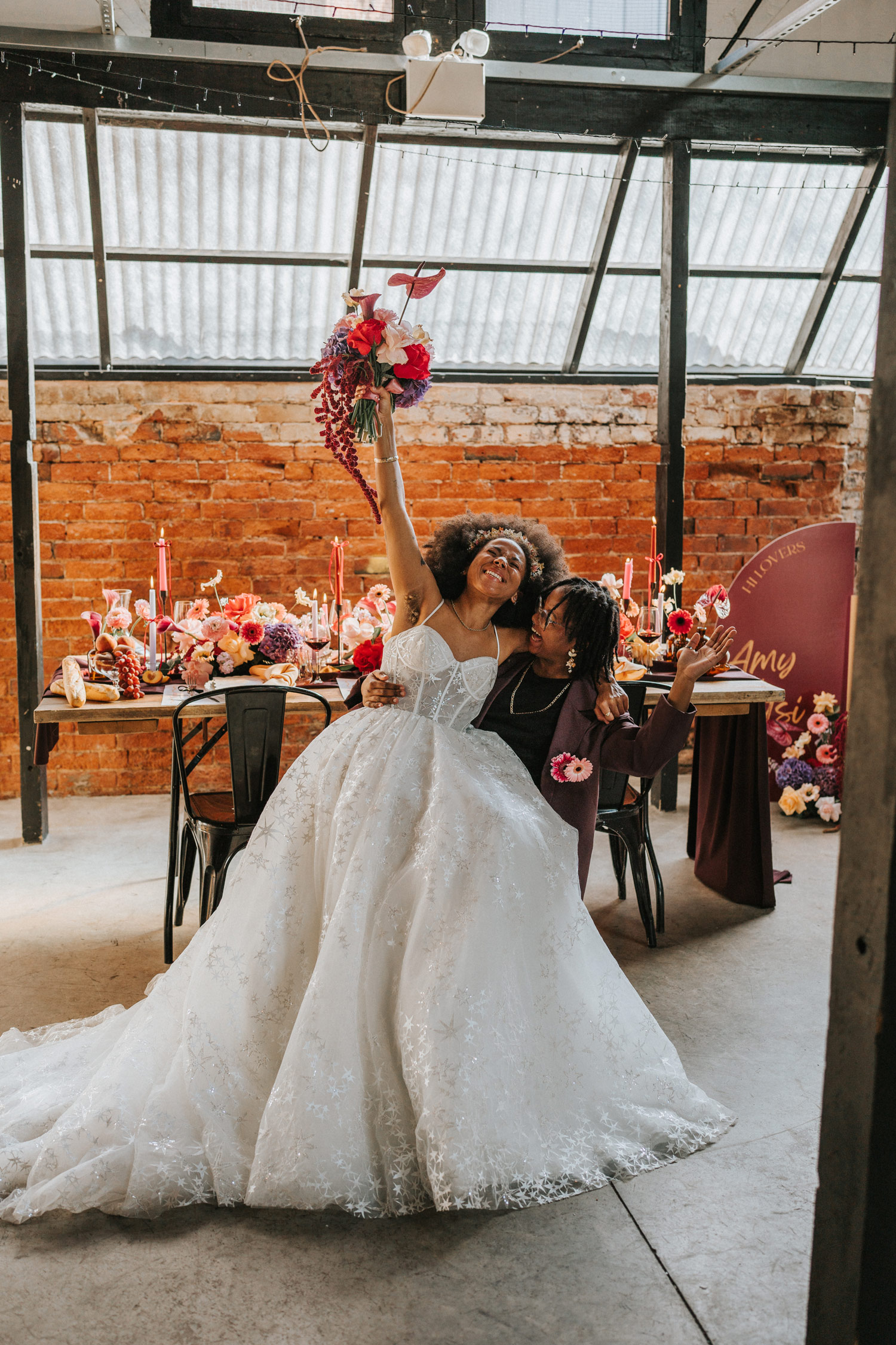 Newlyweds celebrating, one wears a long white wedding gown, holding onto a red bouquet, and sitting on their partner's lap in a barn venue. Taken by Steph Large