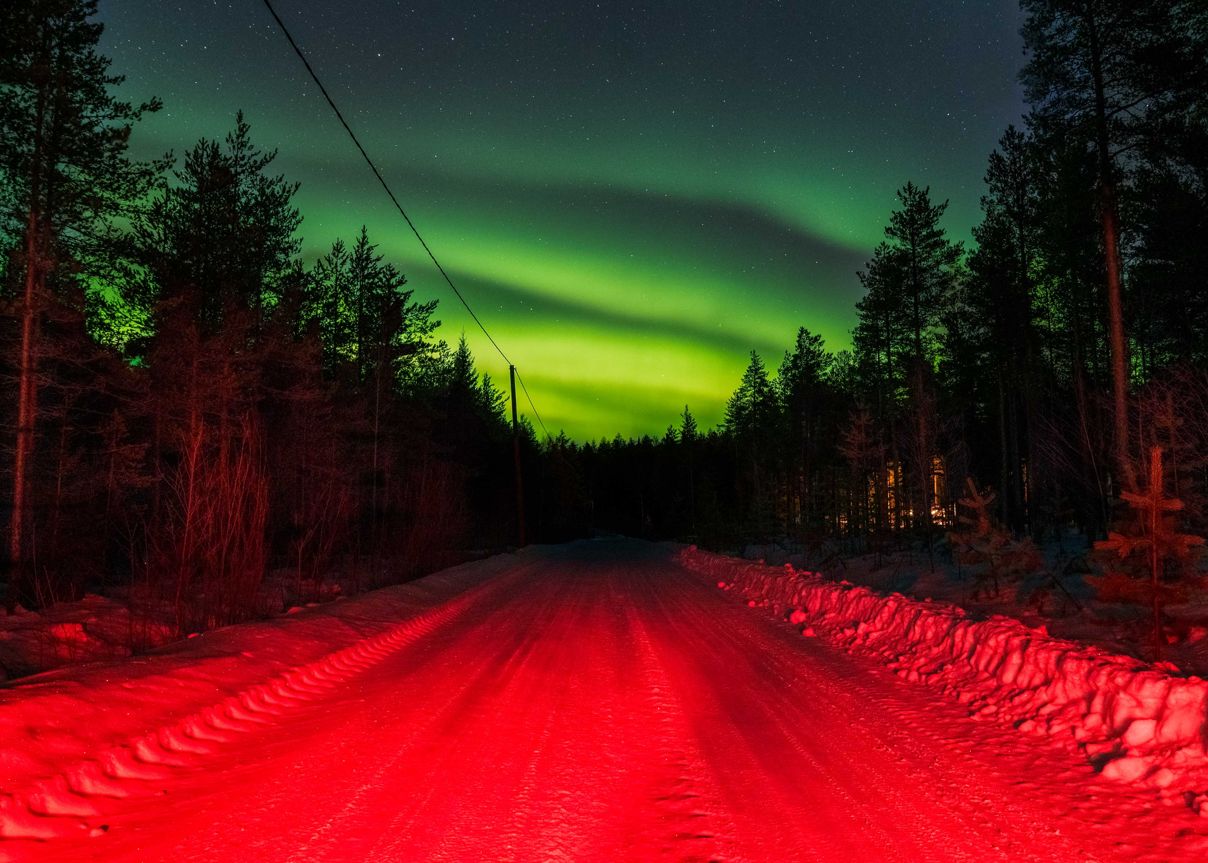 Een weg bedekt met sneeuw verlicht door een sterk rood licht met de groene aurora borealis in de nachtelijke hemel