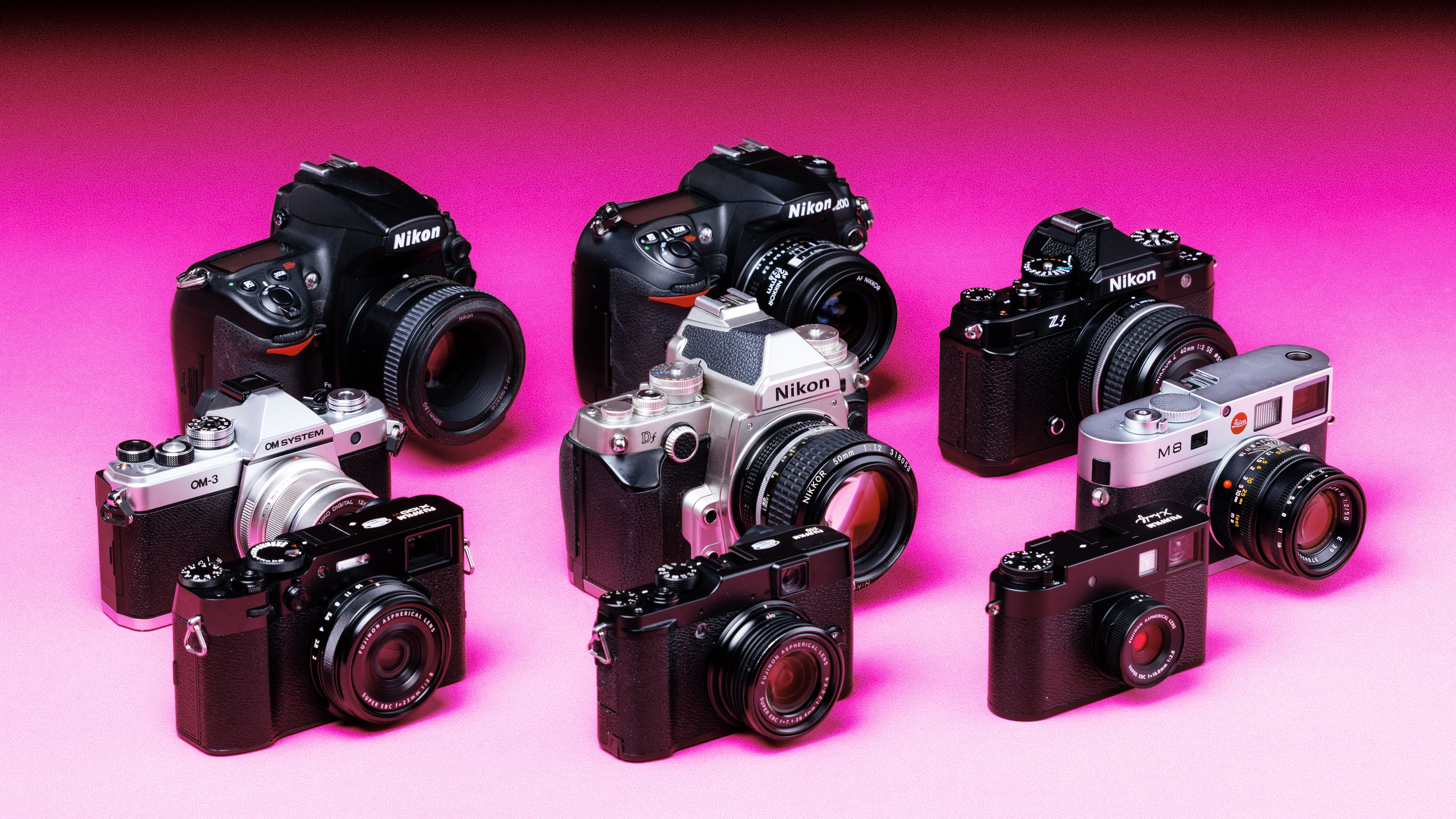 A range of used cameras in a group on a graduated pink background. From left to right, Fujifilm X100V, OM System OM-3, Fujifilm X10, Nikon D700, Fujifilm X Half, Nikon Df, Leica M8, Nikon D200, Nikon Zf
