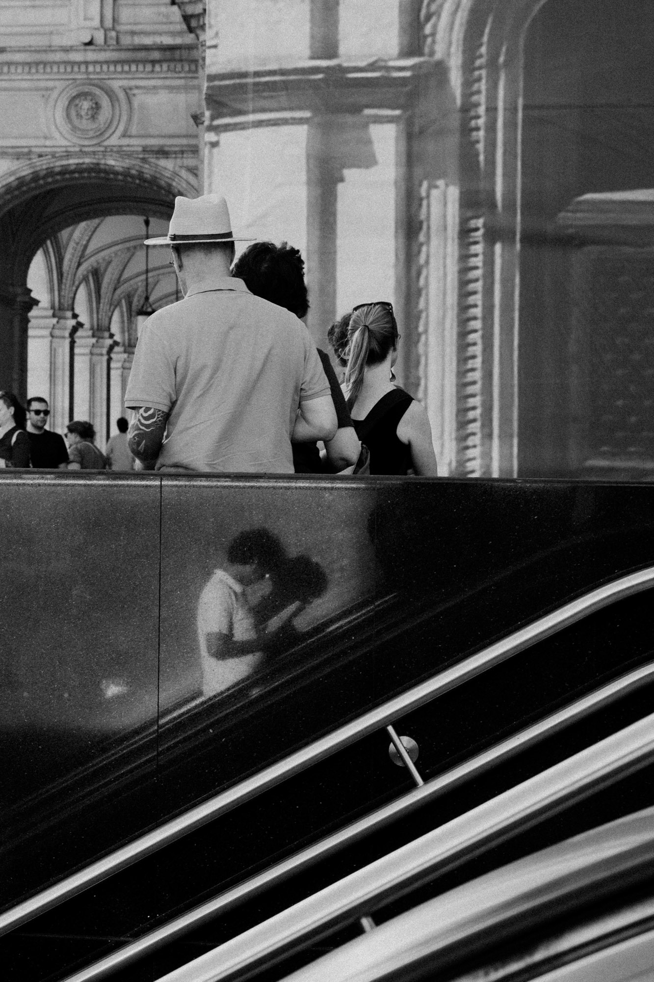 Schwarz-Weiß-Aufnahme einer urbanen Szene mit mehreren Personen vor historischer Architektur. Im unteren Bildbereich verlaufen die Linien einer Rolltreppe. Die Spiegelung einer Person in der Wandverkleidung der Rolltreppe bringt zusätzliche Tiefe in die Komposition. Aufgenommen von Dhan Limwattana mit einer Nikon Z5 II.