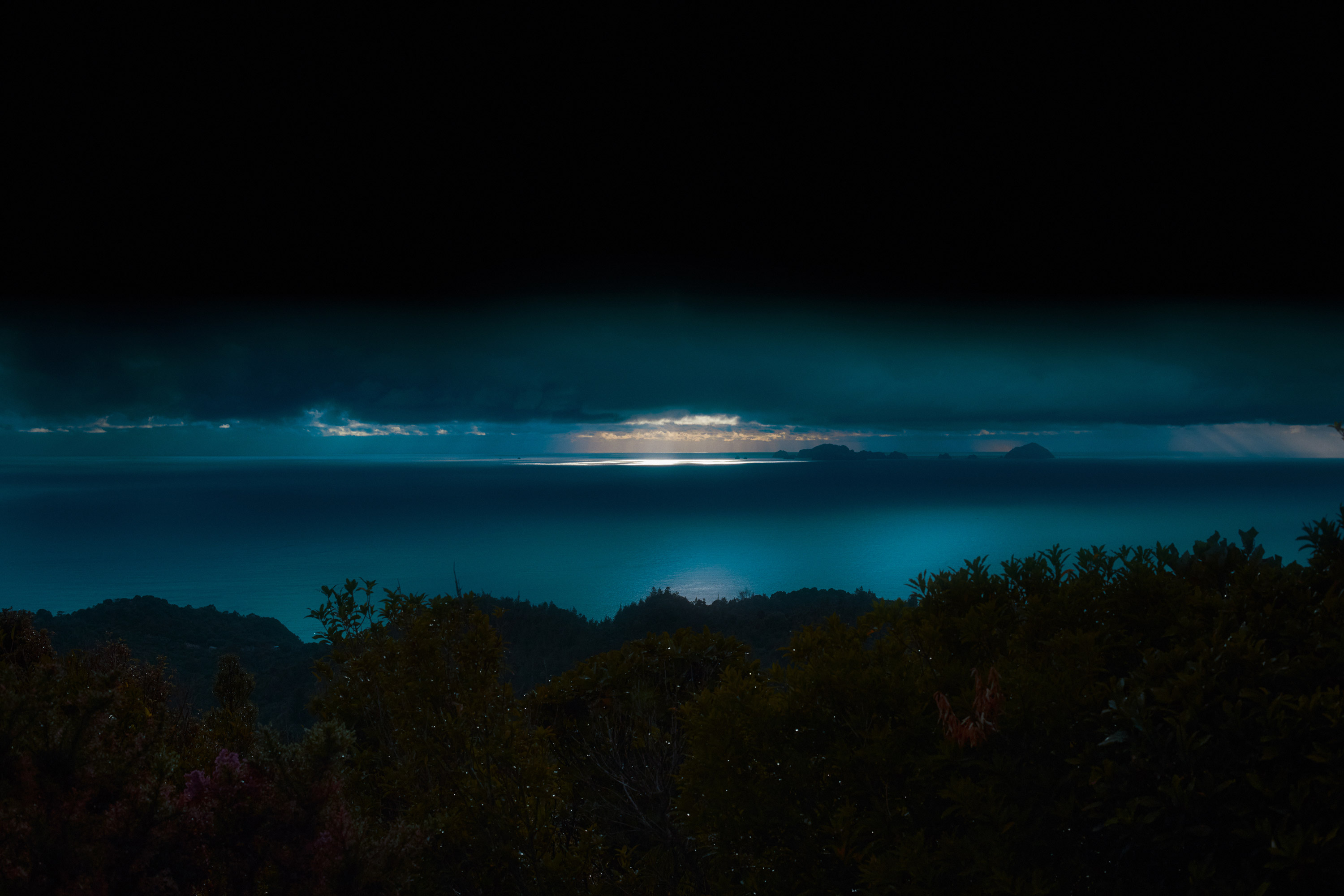 Gewässer in leuchtendem Blau, schwarzes Gebüsch im Vordergrund, Sonnenuntergang im Hintergrund ganz schwach erkennbar, schwarze Wolkendecke, aufgenommen und bearbeitet von Steph O’Connor


