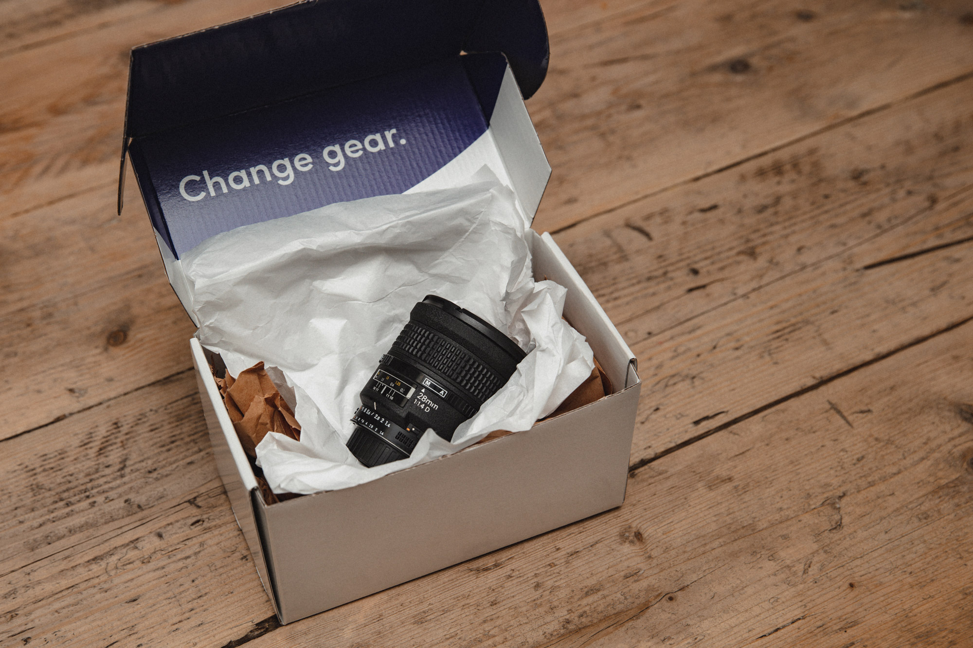 Un objectif 28mm f/1.4 dans un emballage sans plastique MPB sur une table en bois 