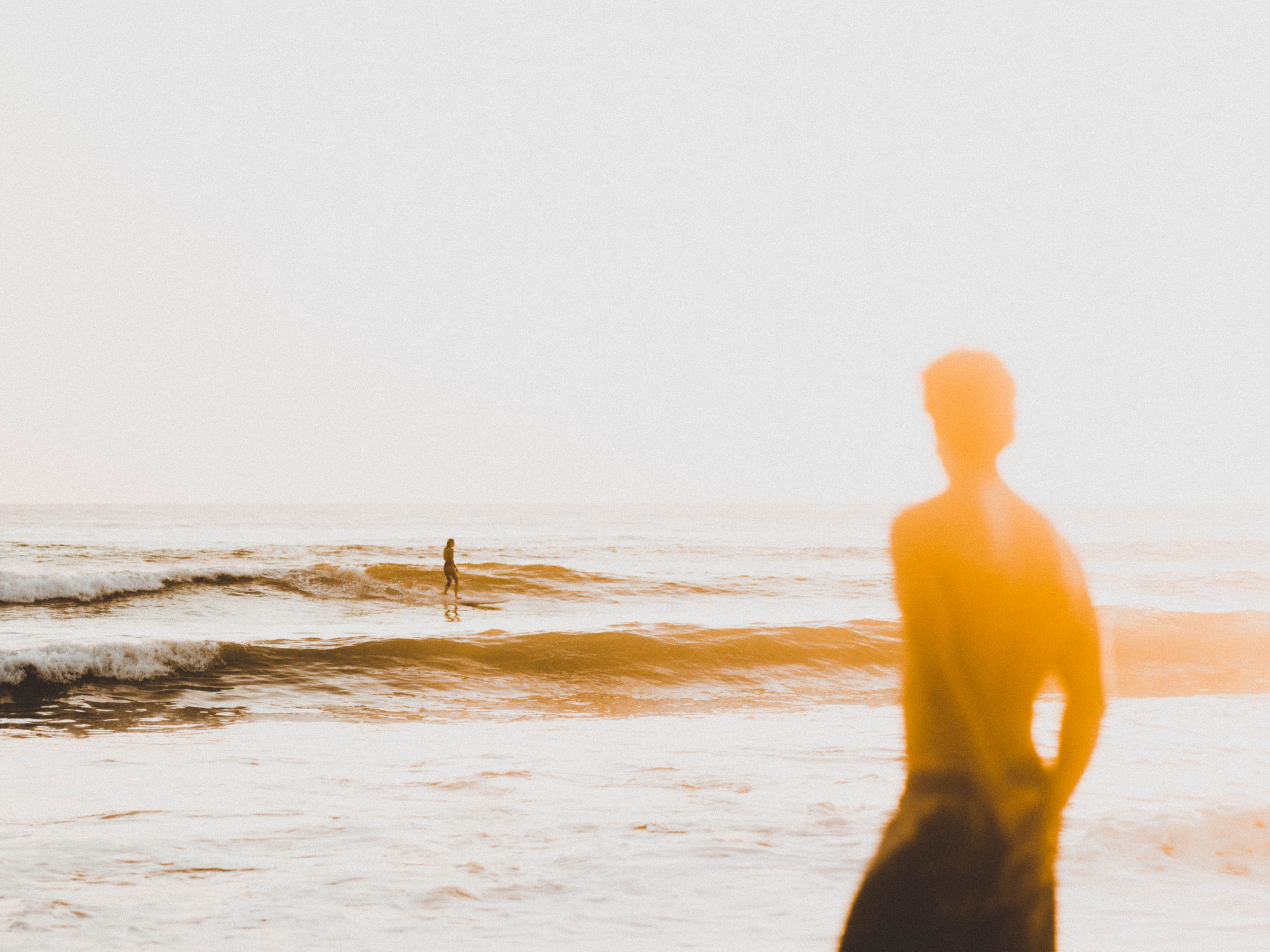 Een persoon op de voorgrond kijkt uit naar een surfer die een golf vangt op zee, gefotografeerd door Ian Howorth met een Hasselblad X1D II middenformaat camera