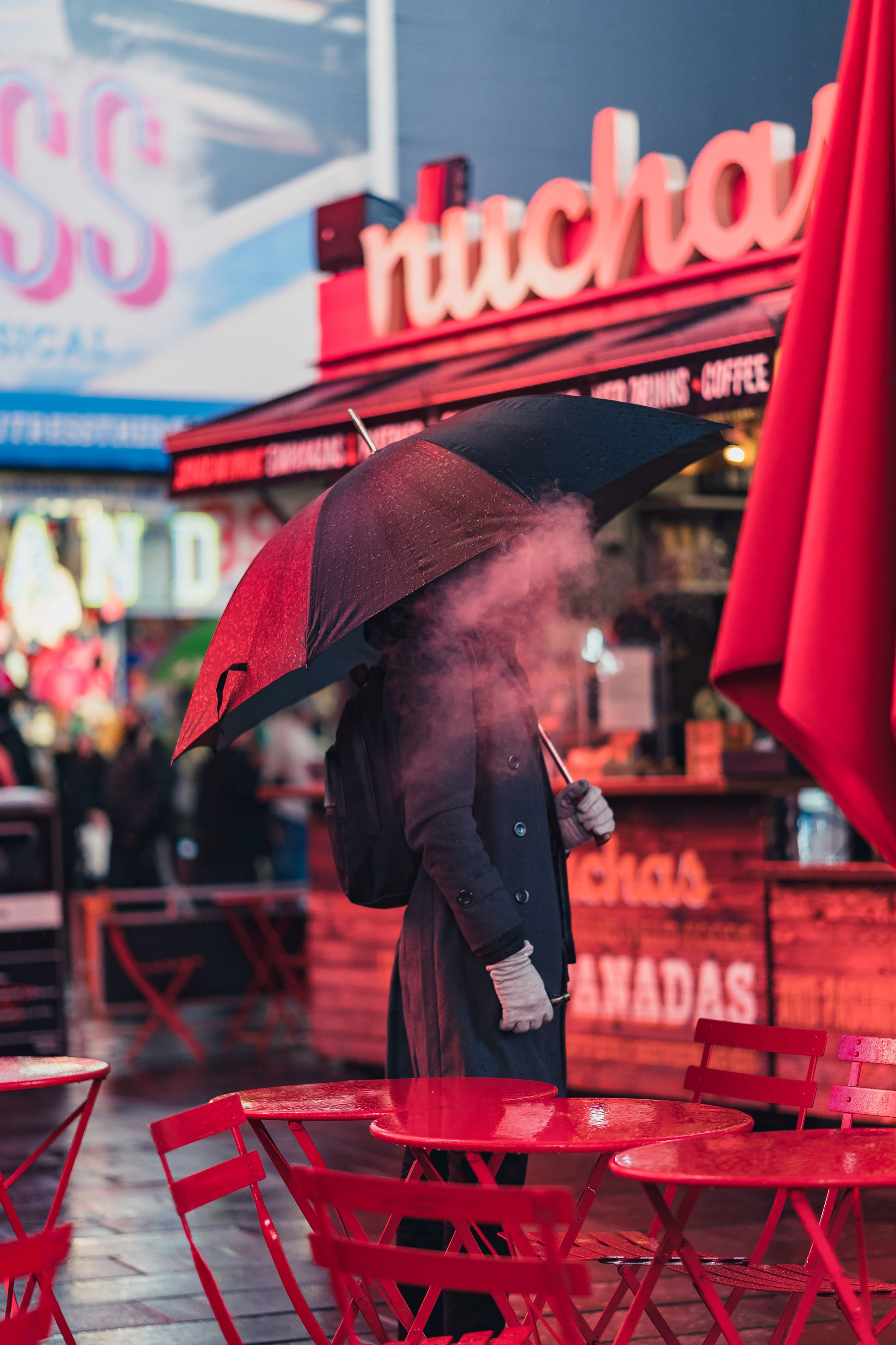 Person at night holding an umbrella with a cloud of smoke obscuring face in front of red signs and tables in Times Square in New York City. Photo by Manuel Pena on a Sony A7 III.