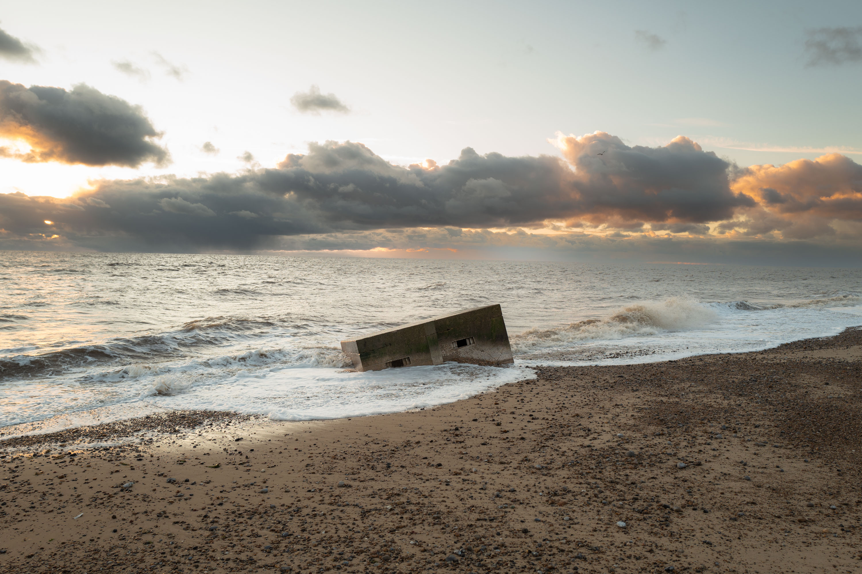 An old pillbox lays on its side in the middle of a beach as waves crash into it. By Ian Howorth.