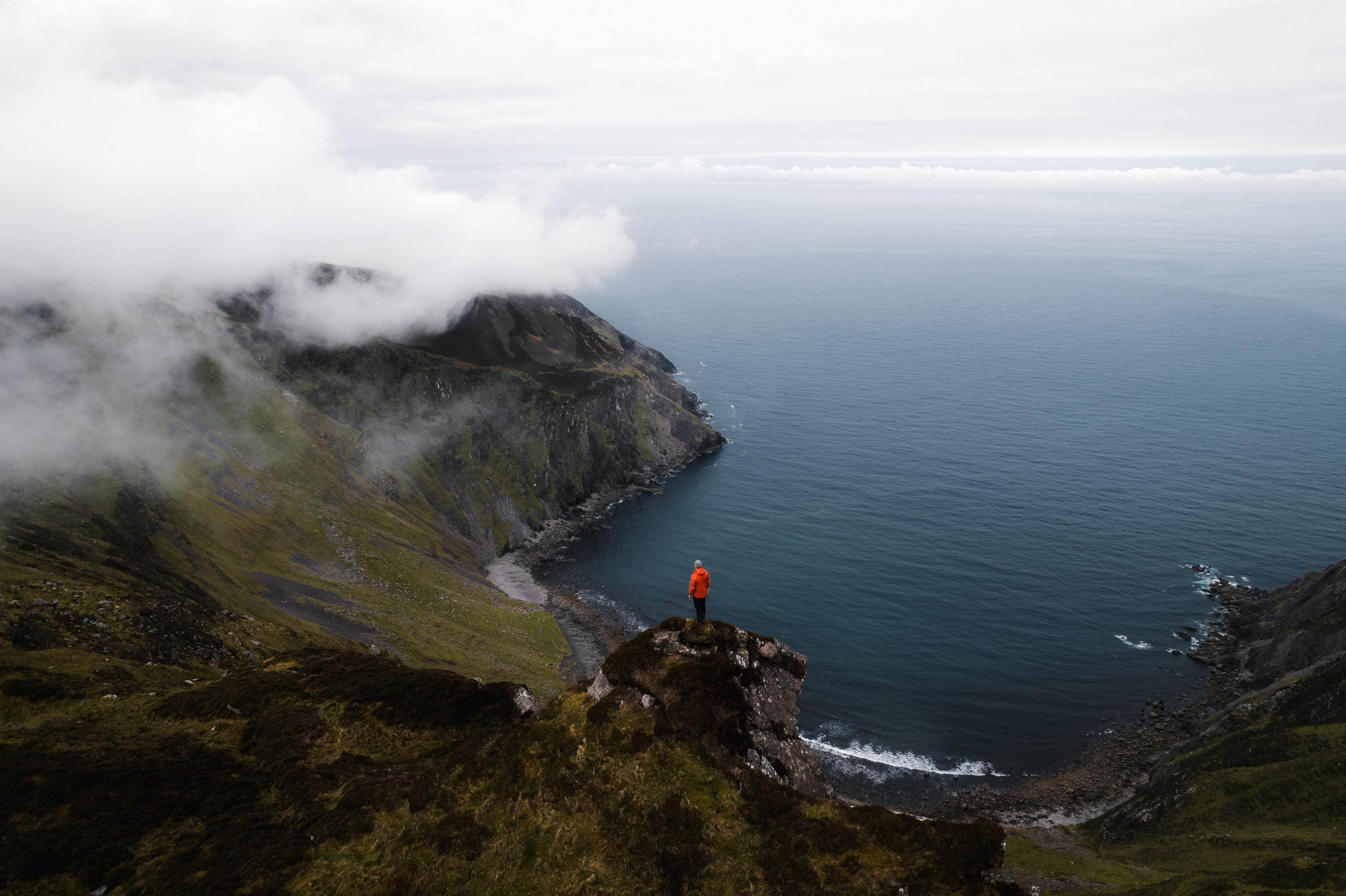 Drohnenaufname einer wolkenverhangenen Klippenlandschaft, auf der ein Mensch in roter Jacke zu sehen ist.