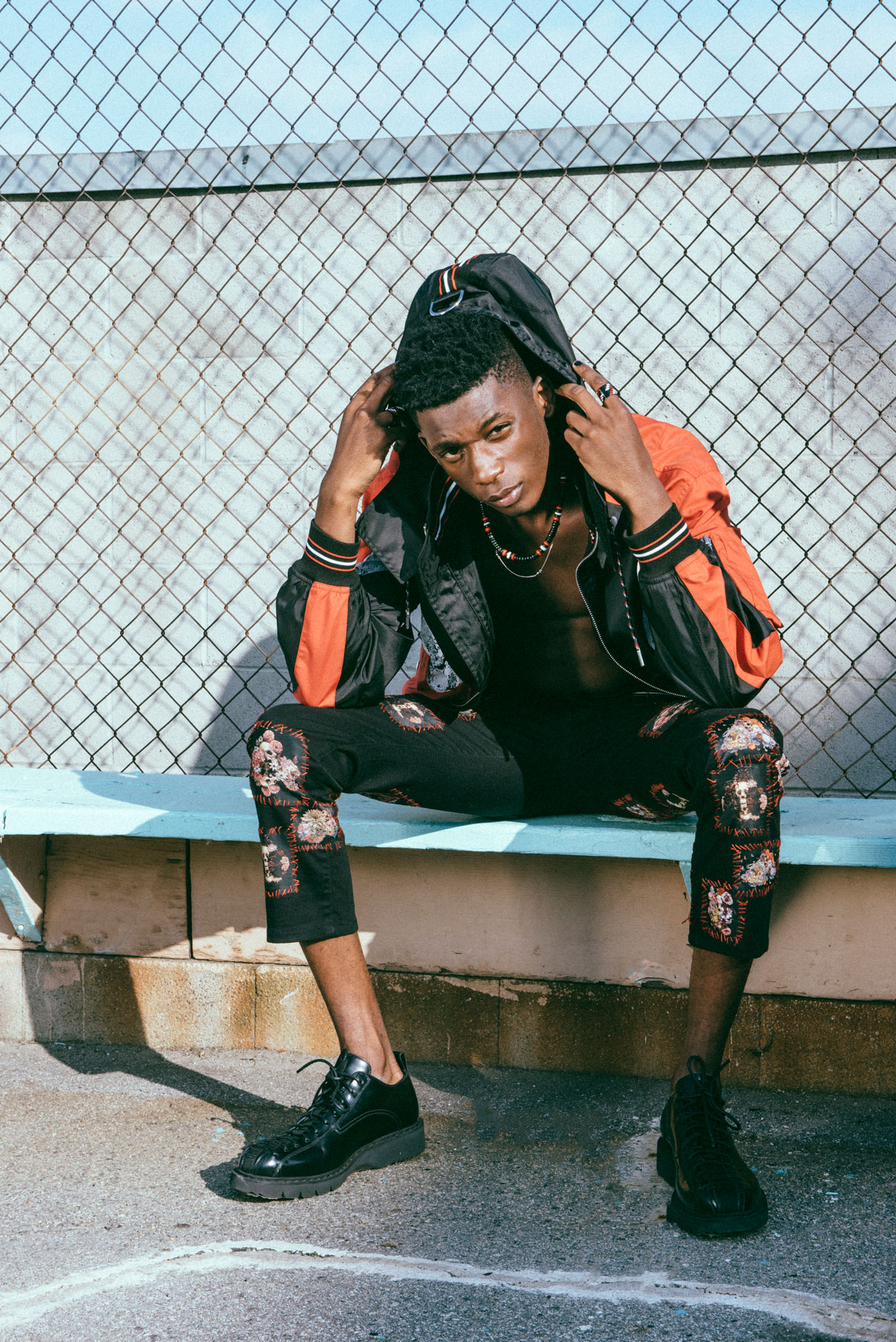 Image of a man in black and orange jacket sitting on a blue bench during an editorial photoshoot by Lindsey Byrnes.