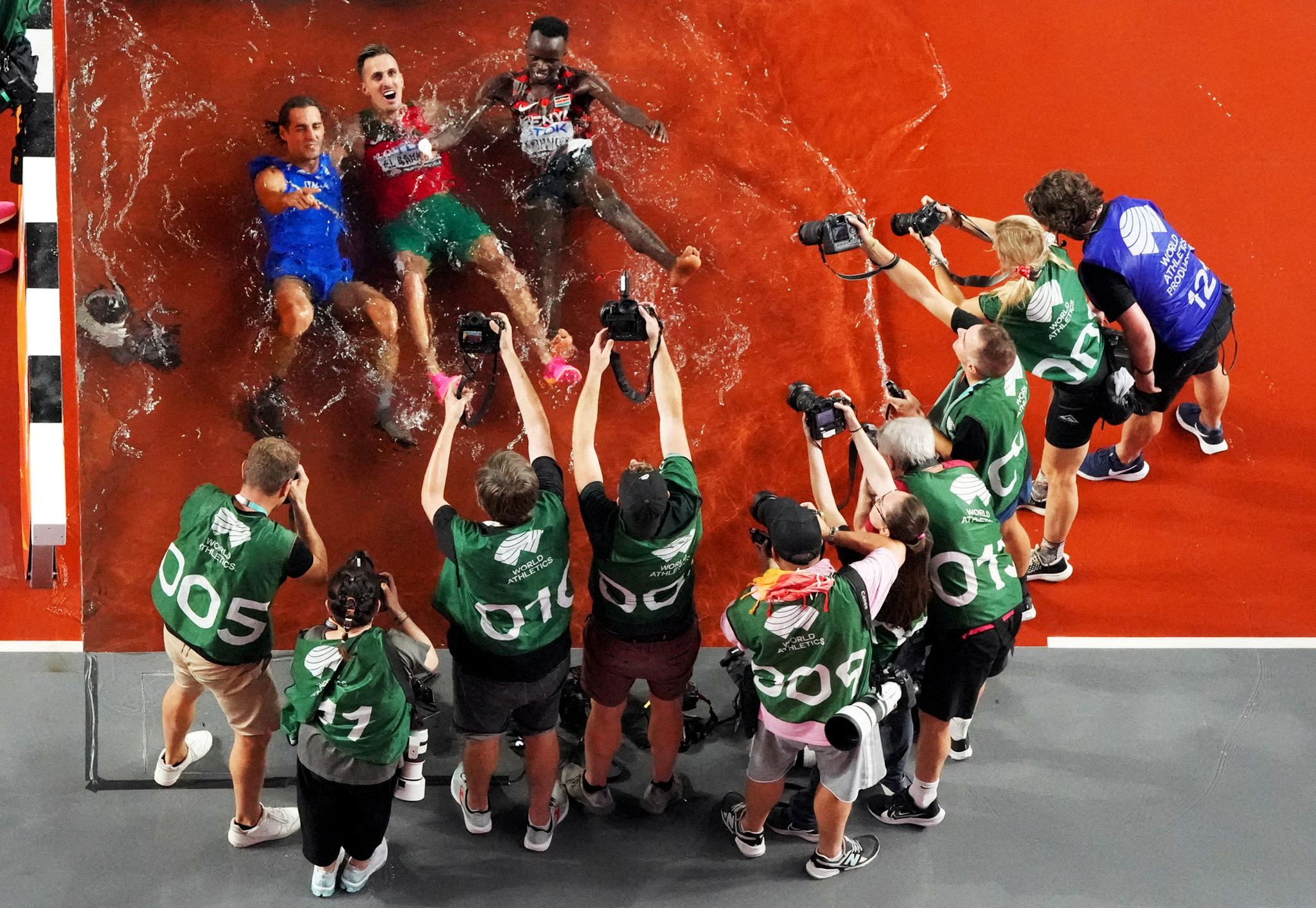 A top down drone shot of photographers taking pictures of three athletes who are celebrating in a pool of water in an athletics field. By Fabrizio Bensch.