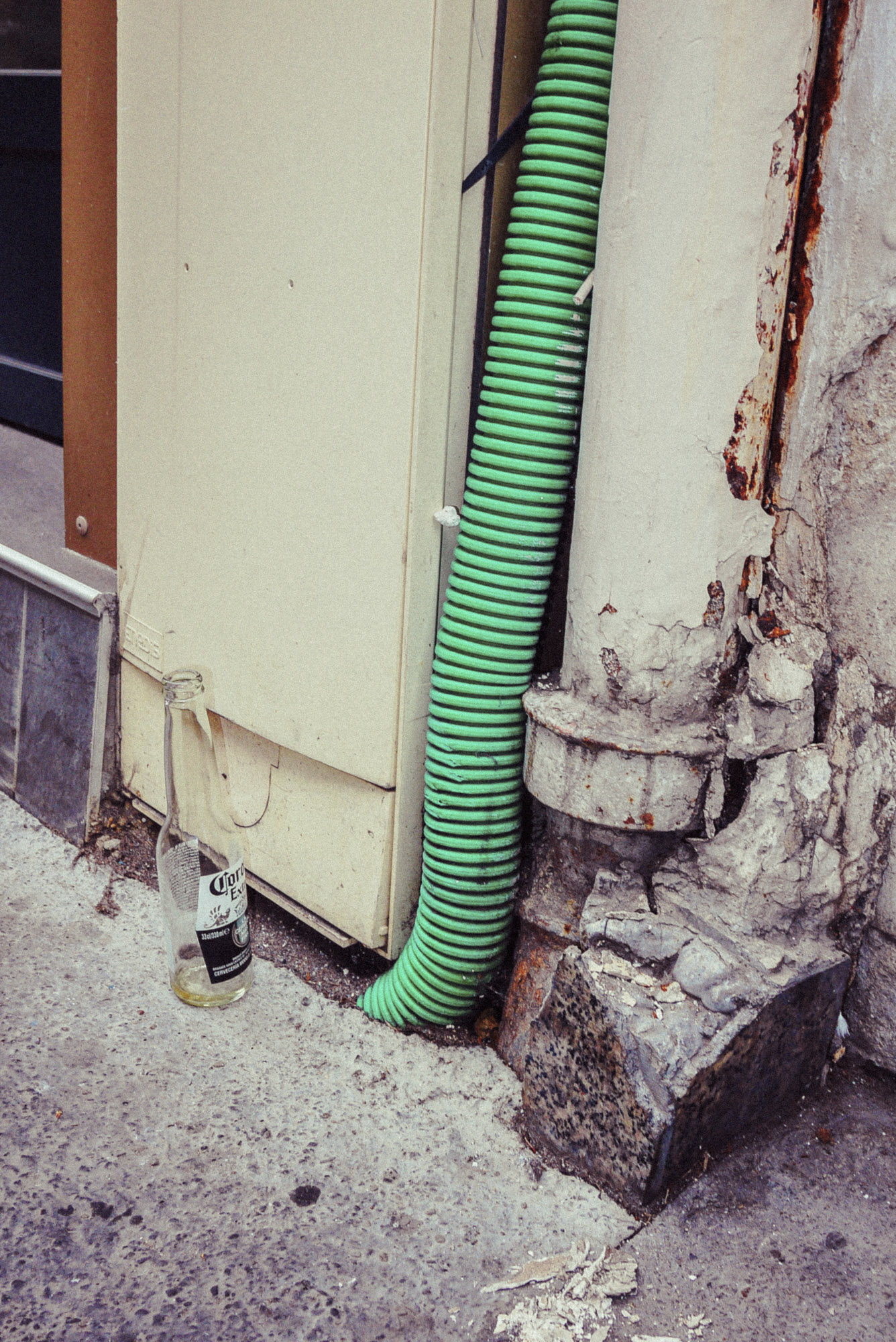 Streetaufnahme mit einem grünen Abflussrohr, das an einer abgenutzten Pariser Hauswand hinunterläuft. Davor eine Glasflasche, die auf dem Gehweg neben abblätternder Farbe und rauen Texturen steht. Aufgenommen von Wesley Verhoeve mit einer Panasonic Lumix GF1.