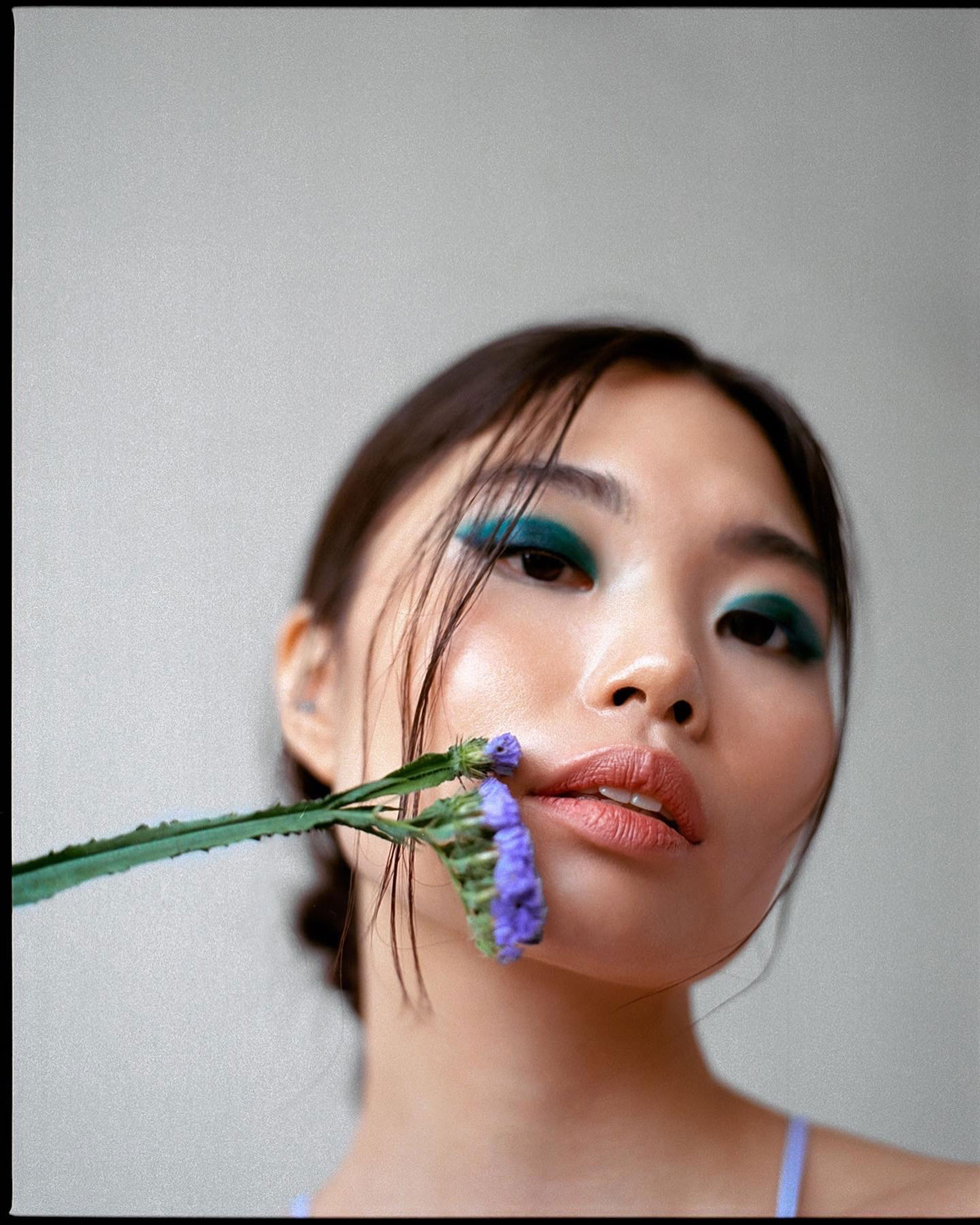 Portrait of a woman with turquoise eyeshadow and purple flowers next to her mouth, against a grey background, shot by Samia Rachel
