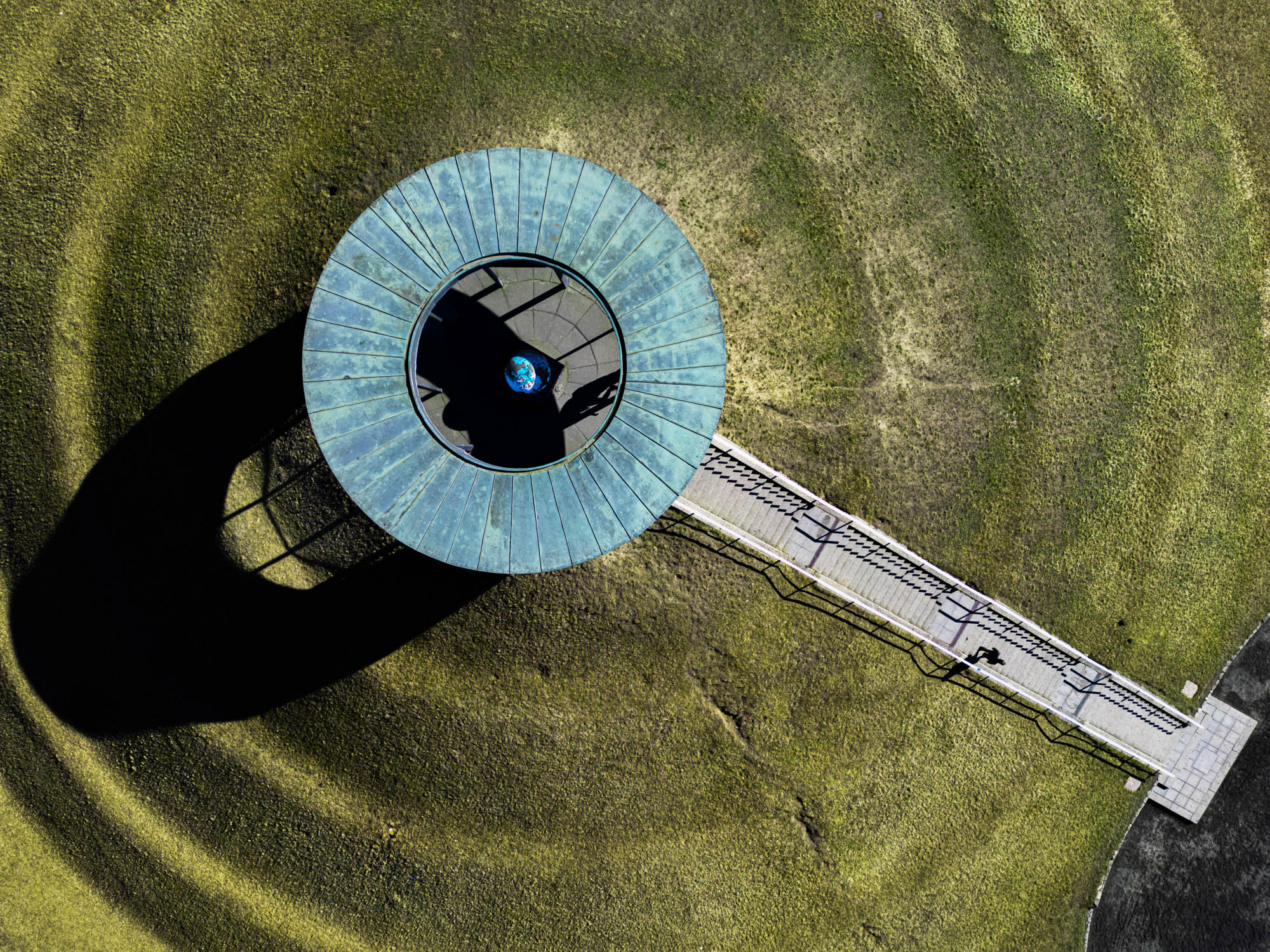 Senkrechte Luftaufnahme des Lilienthal-Denkmals auf dem Berliner Fliegeberg mit blaugrünem Dach, langer Treppe und markantem Schatten auf der umliegenden Wiese. Aufgenommen von Joel Arias mit einer DJI Mini 3 Pro. Aufgenommen von Joel Arias mit einer DJI Mini 3 Pro.