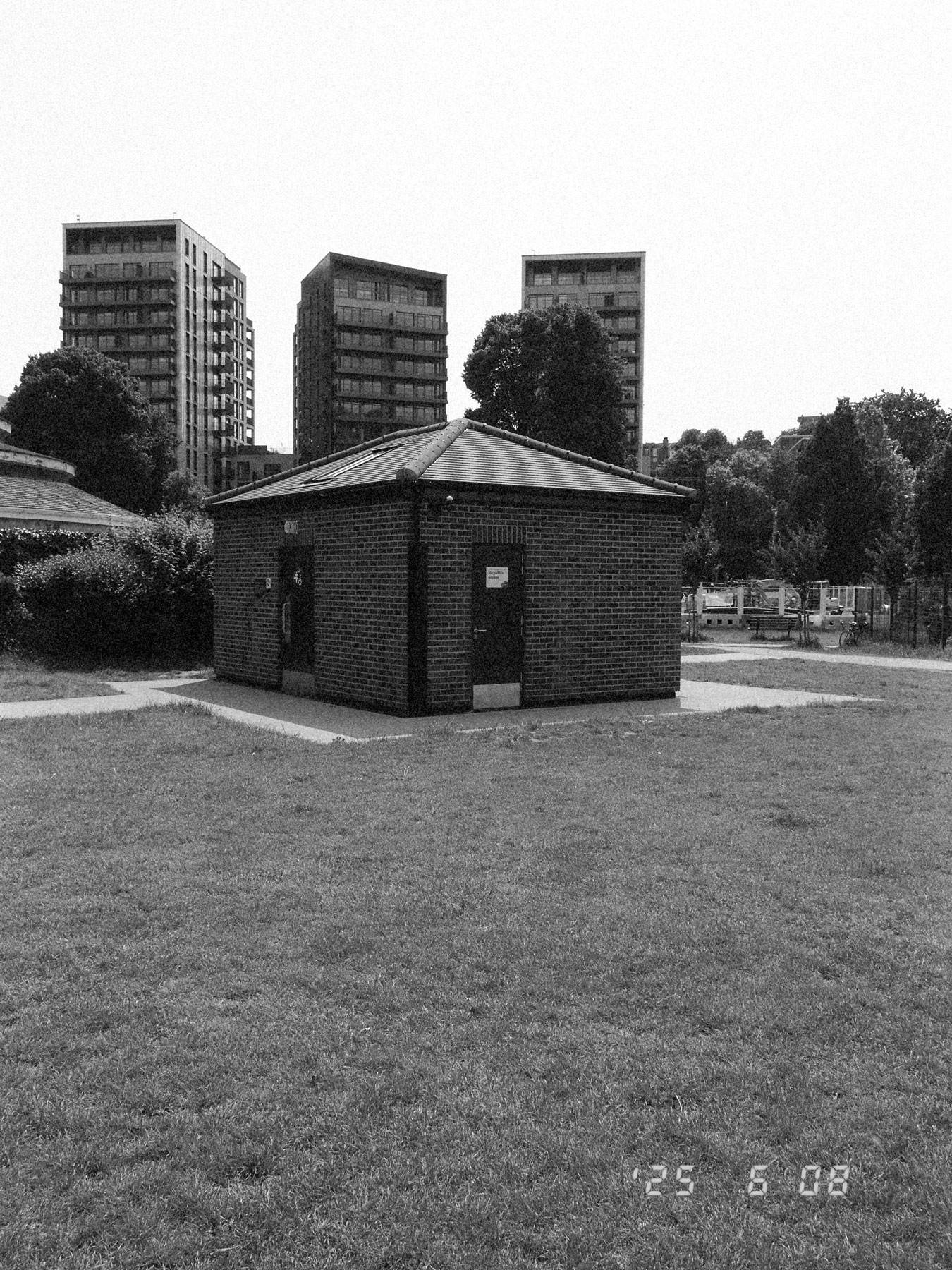 Schwarz-Weiß-Aufnahme eines kleinen Gebäudes in einem Park mit drei Hochhäusern im Hintergrund.