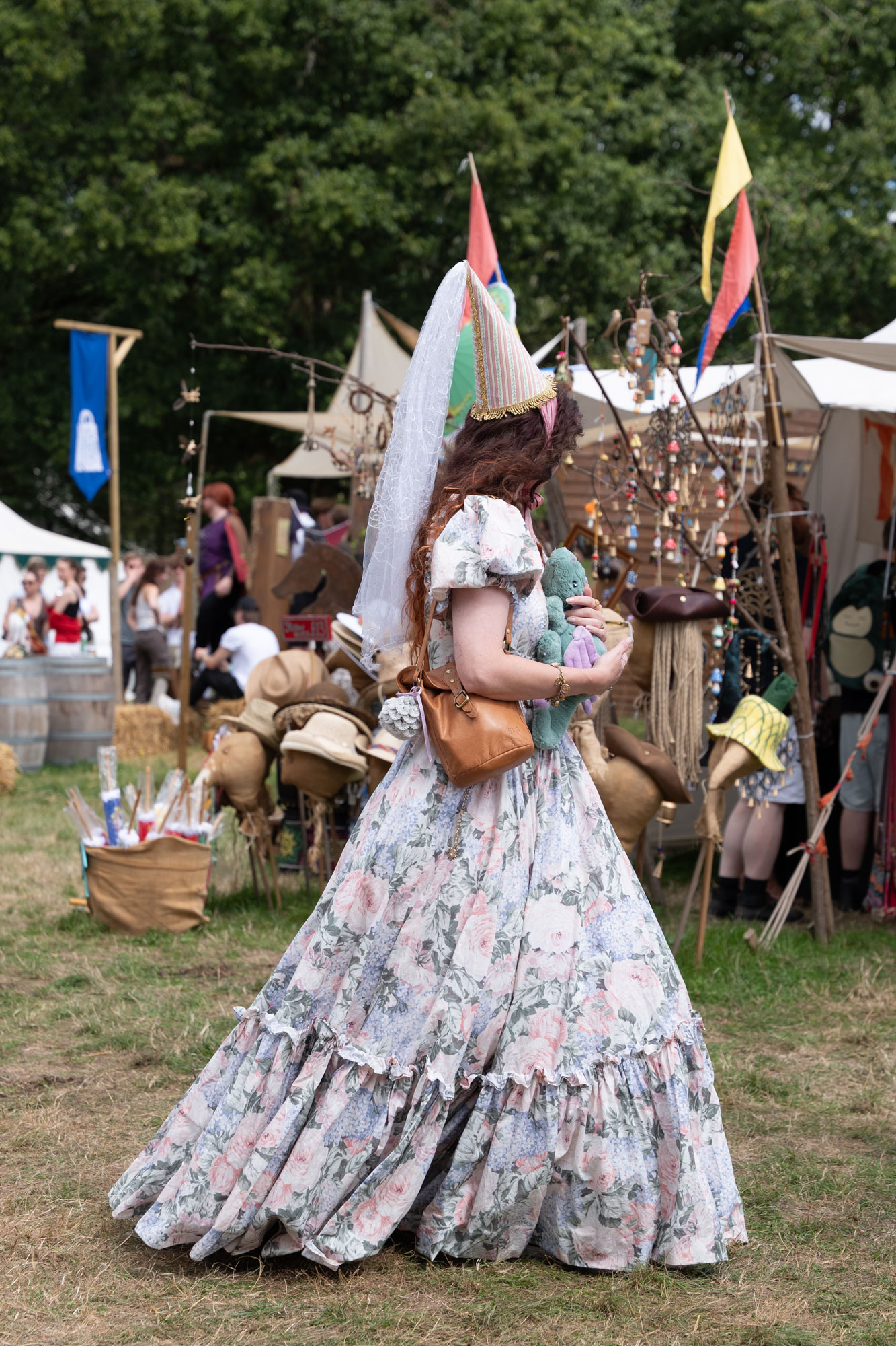 A person with long curly hair dressed in a flowing, floral medieval gown and a pointed hat. They are holding a stuffed toy. Stalls selling various medieval trinkets and costumes are in the background.