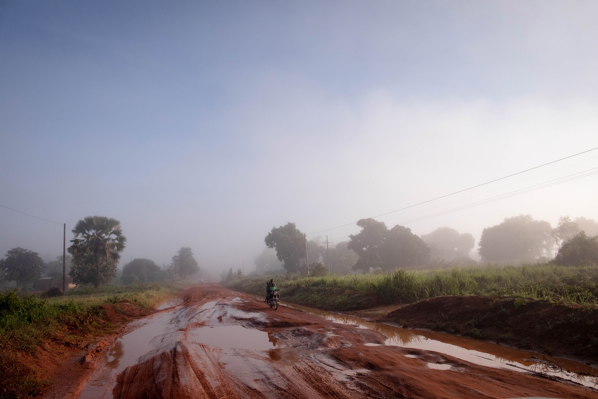 Une route boueuse en Ouganda, avec des arbres en arrière-plan et un cyclomoteur roulant sur la route, photo prise par Thomas Williams avec un Canon EOS 5D Mark IV.