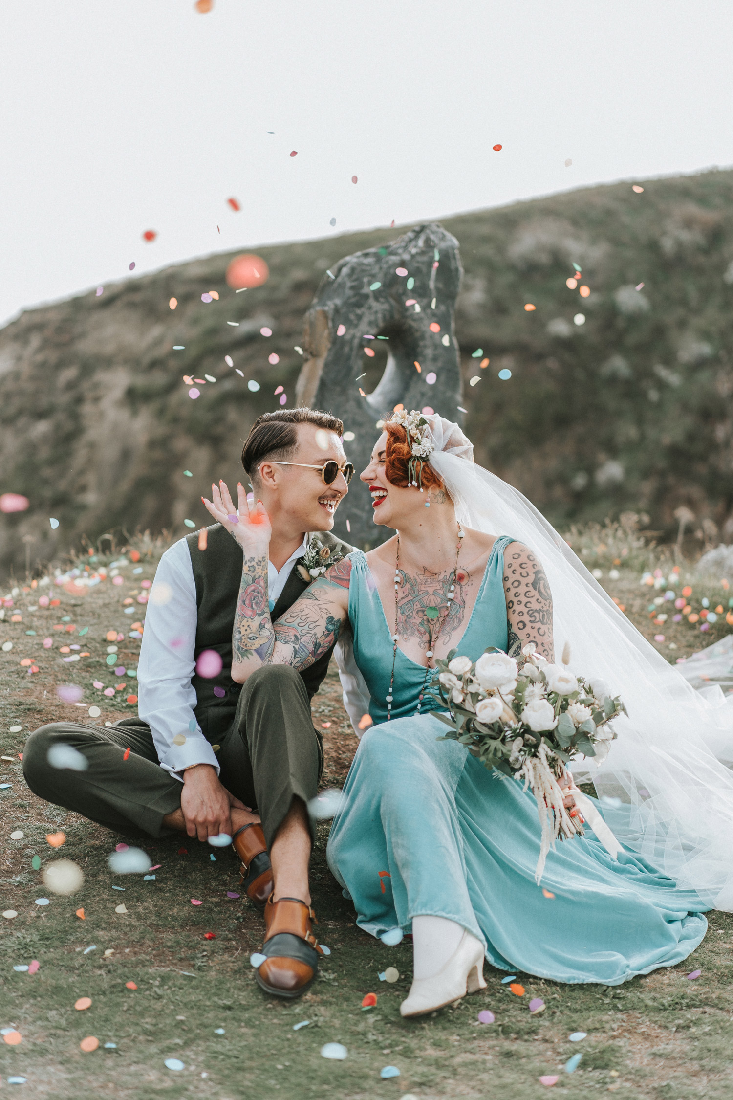 Newlyweds Max and Lori smiling and celebrating with confetti, sitting on a grassy hill. Lori wearing an aqua dress and holding a white bouquet of flowers. Shot by Steph Large.
