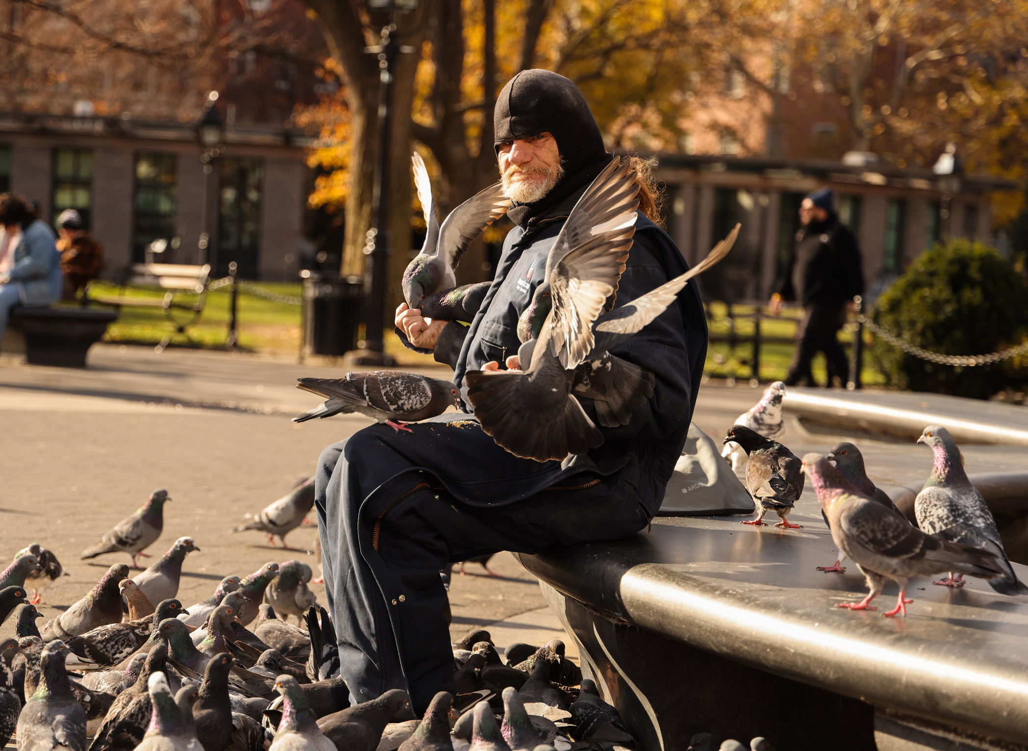 Man in New York City’s Washington Square Park sitting on a bench feeding a group of pigeons. Taken with Canon EOS R5 by Berta Amelinaite.
