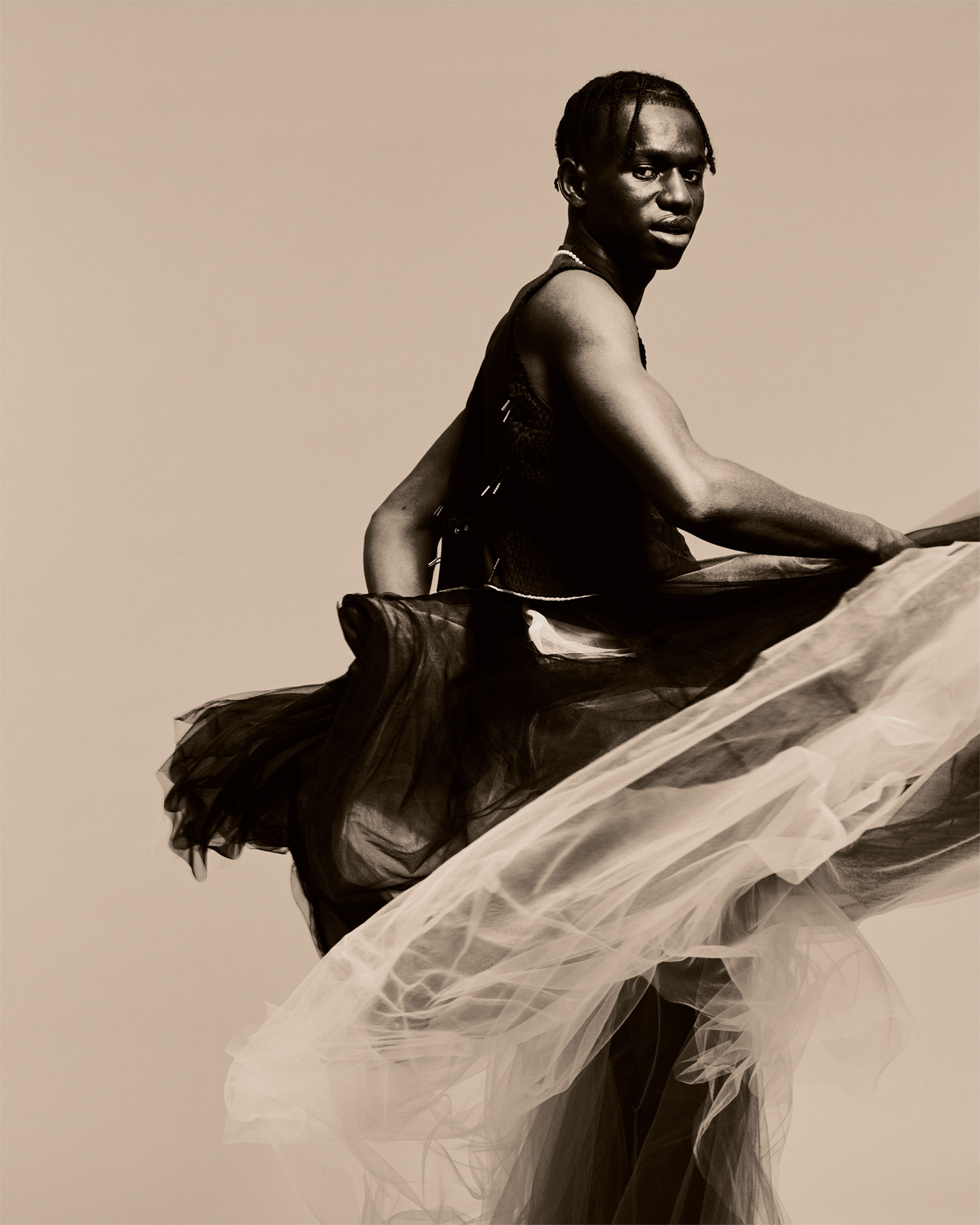 Black man with billowing fabrics in black and white, which he swirls around him, against a sand-coloured background, shot by Lisa Marie Asubonteng