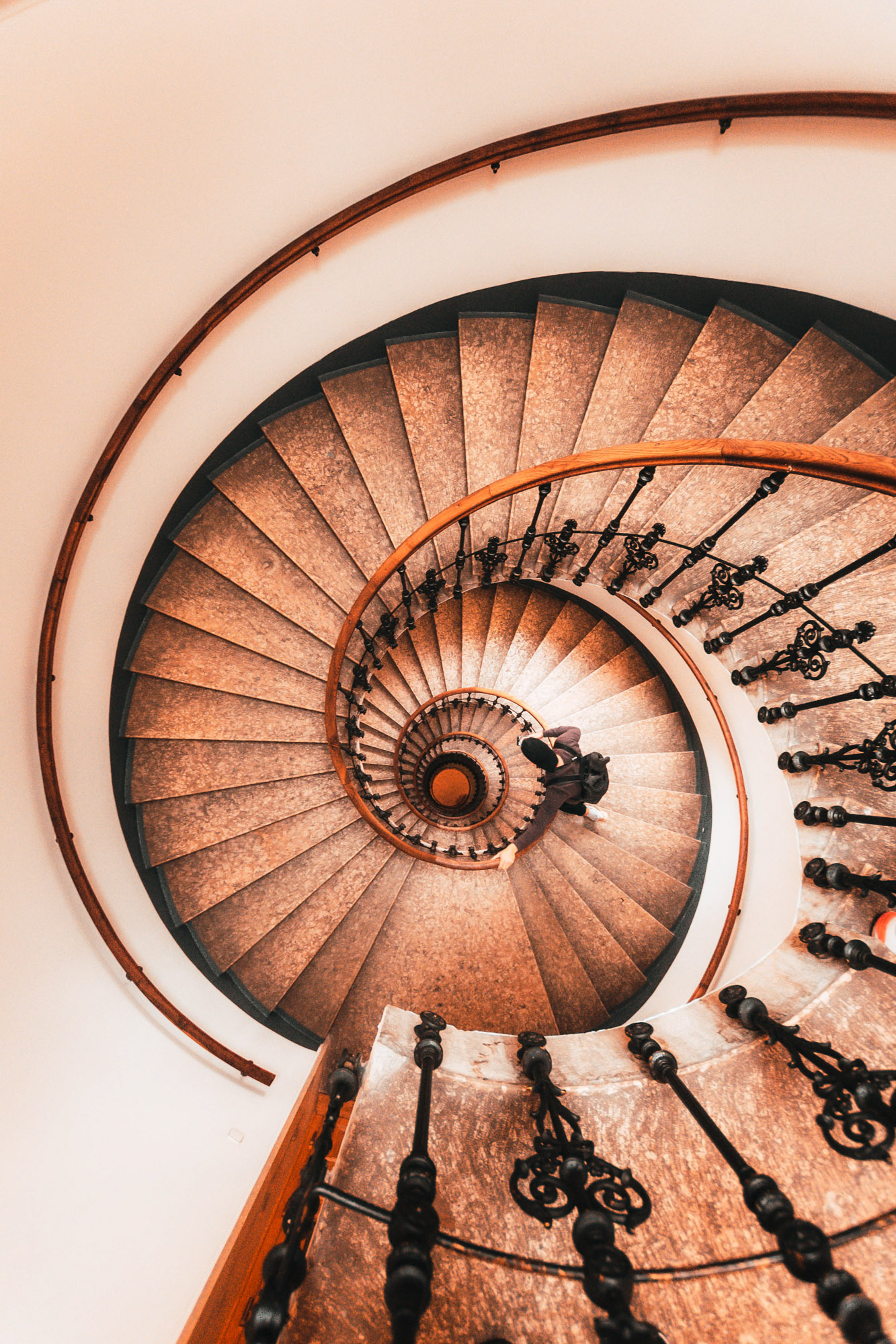 Blick von oben auf eine große Wendeltreppe mit hölzernem Handlauf. Die Stufen bilden eine Spiralform und unten läuft eine Person. Aufgenommen von Dhan Limwattana.