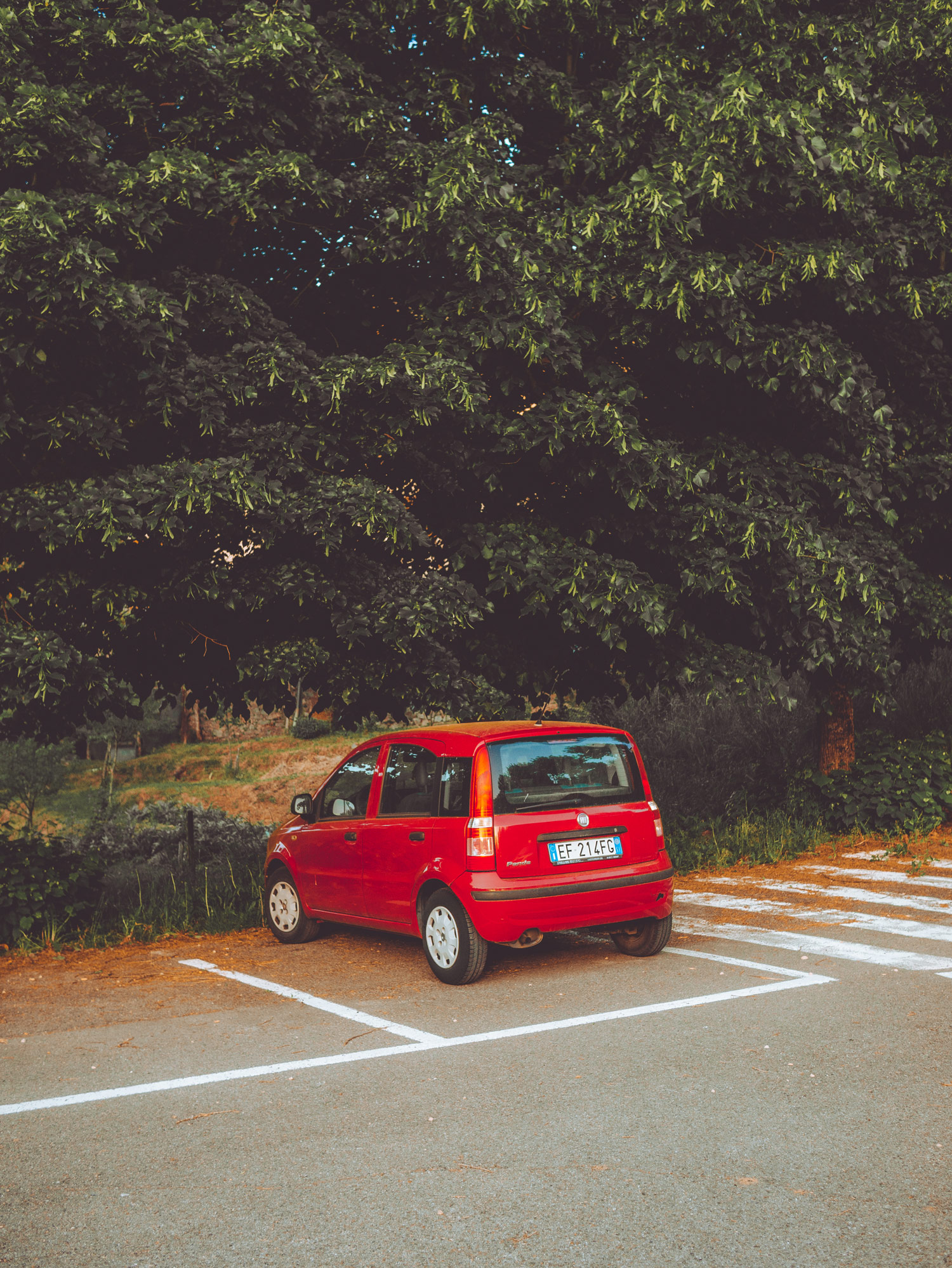 A red car parked in a car park in Petrolo, Tuscany. There are green trees in the background.