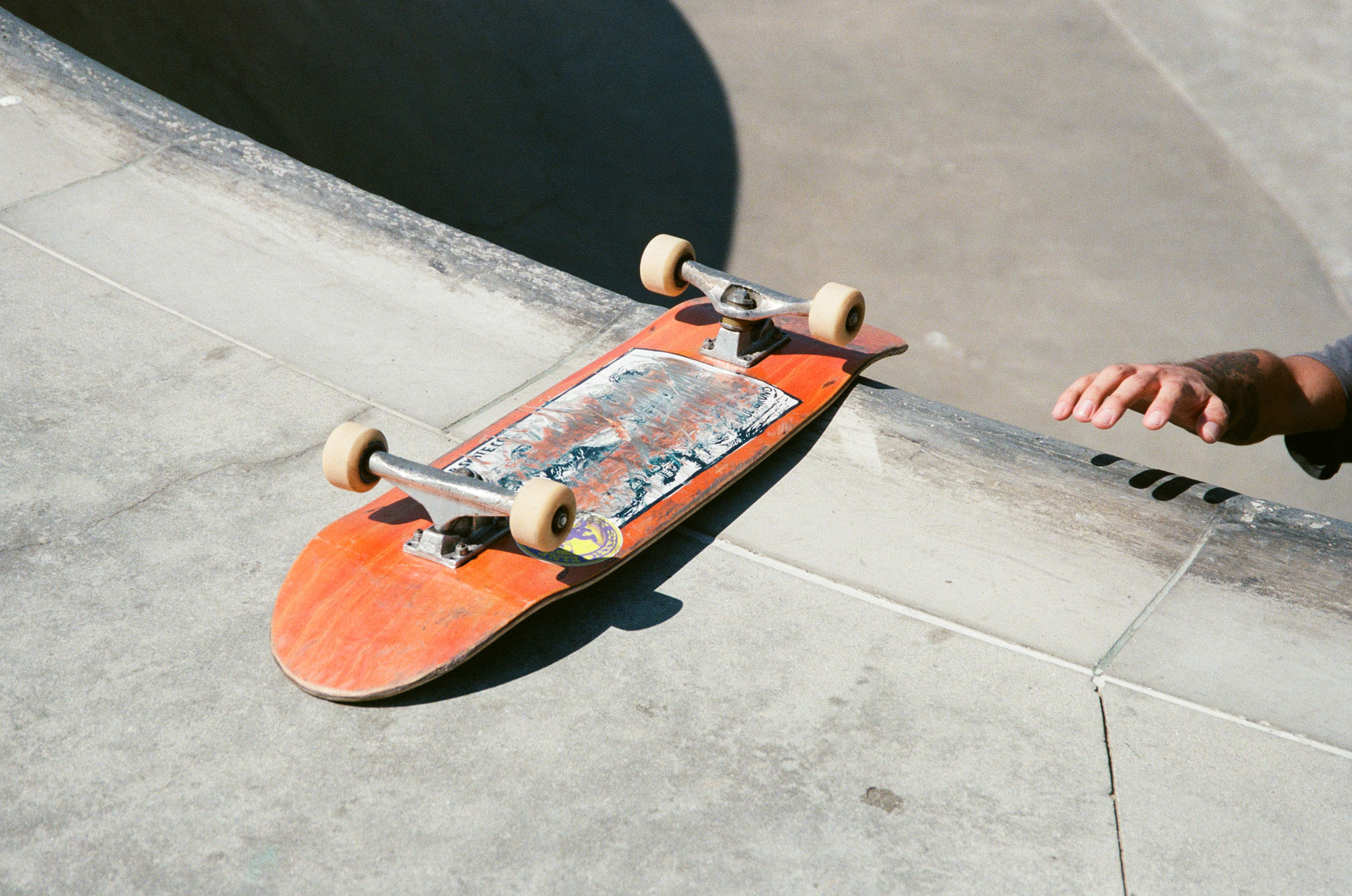 Hand greift nach orangefarbenem Skateboard im California Skatepark.