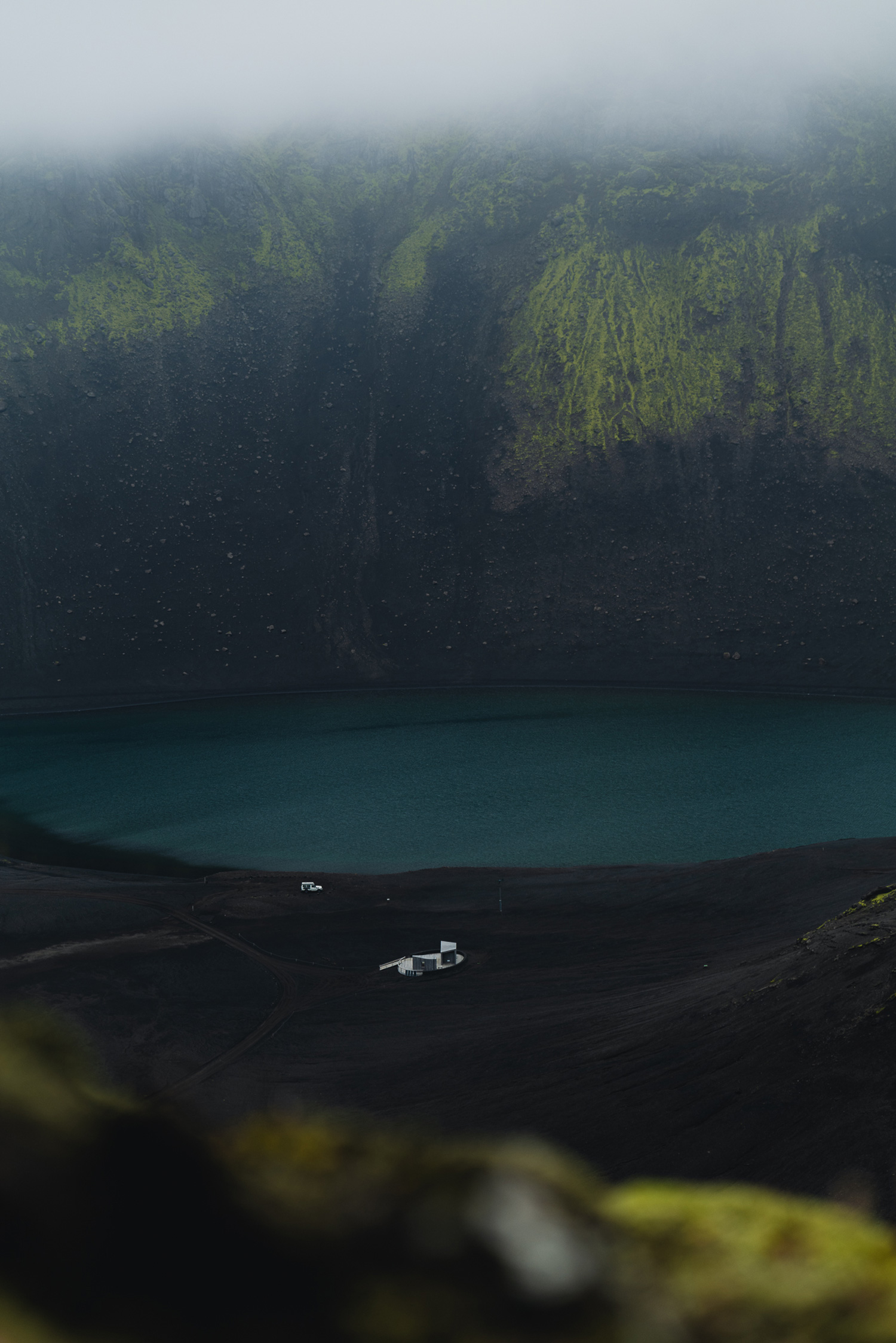 A lake in Iceland appears at the bottom of a mountain range covered in grass, with a white building and a car next to the river, shot by Paul Watson with a Panasonic S1R