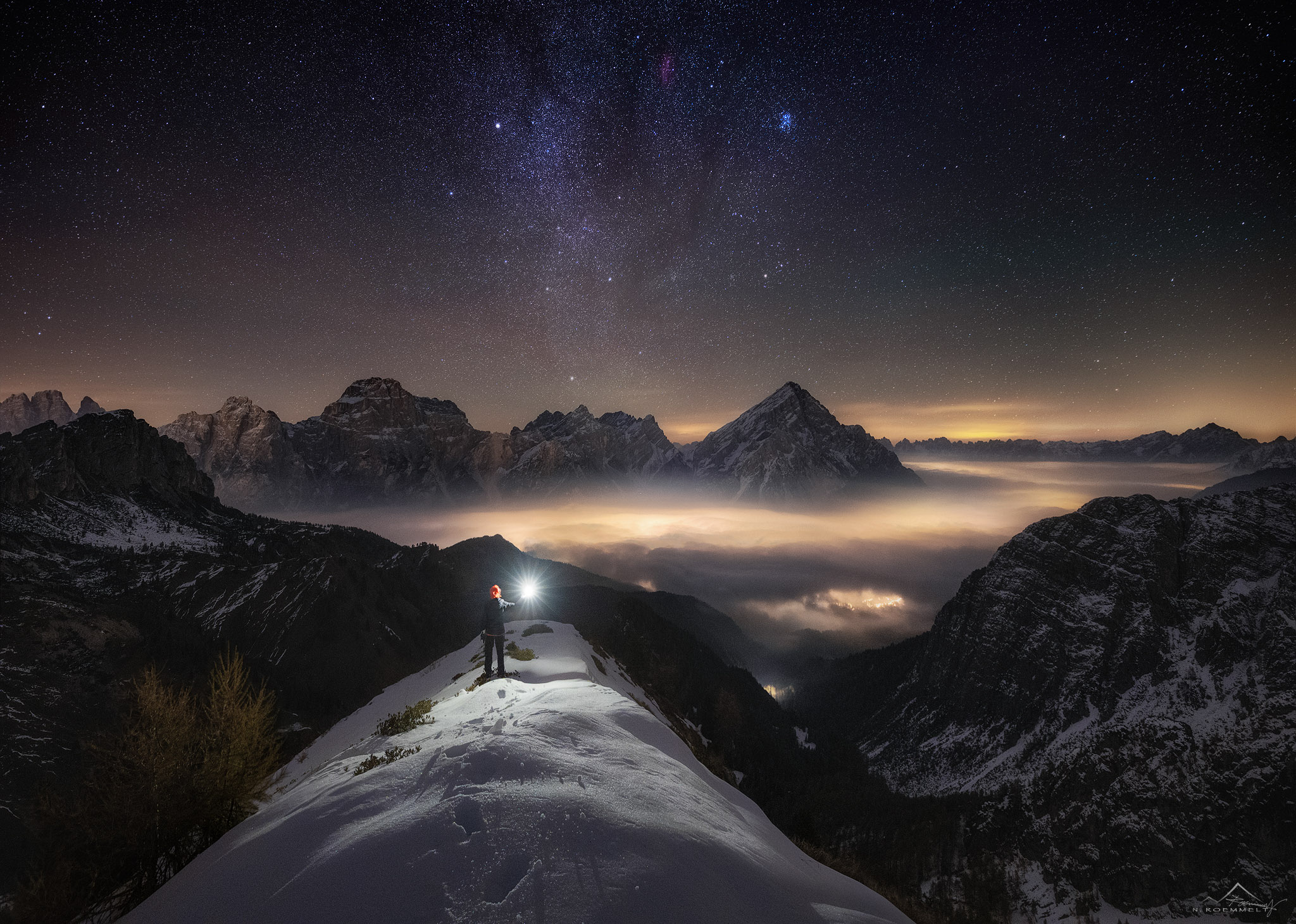 Langzeitbelichtung auf einem Berggipfel mit einer Person, die eine Taschenlampe nach oben hält. In der Ferne sind die Lichter der Stadt, die Sterne und die Galaxie zu sehen.