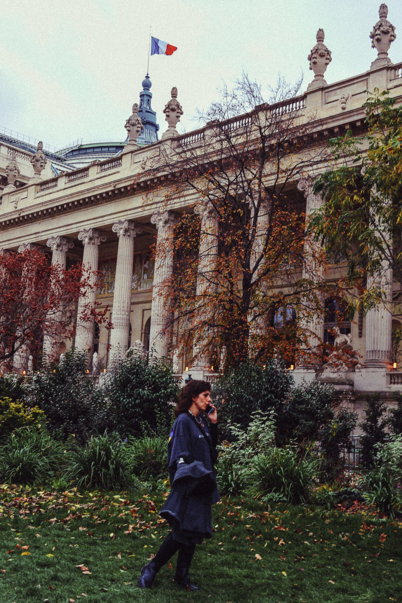 Streetaufnahme einer Person, die telefonierend in einem Pariser Park mit Herbstlaub vor einem historischen Gebäude mit Säulen steht, auf dem die französische Flagge weht. Aufgenommen von Wesley Verhoeve mit einer Panasonic Lumix GF1.